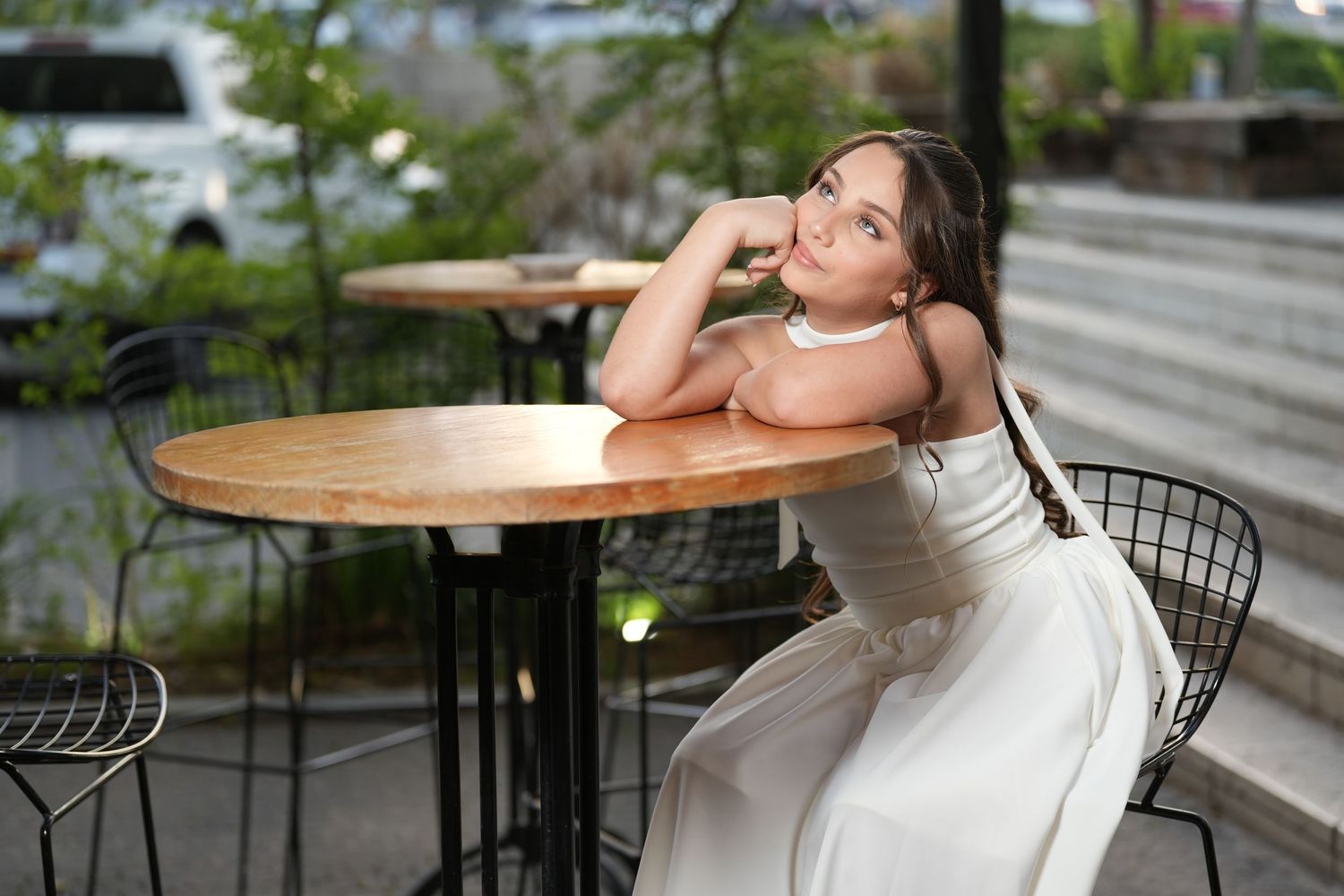 A young woman in a white dress sits gracefully at an outdoor cafe table, resting her chin on her hand.