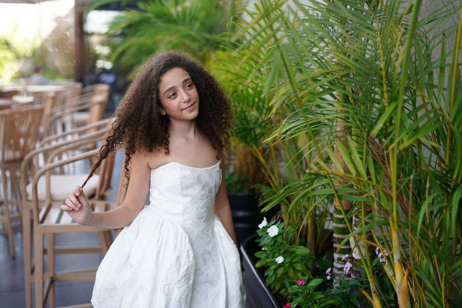 Woman in flowing white dress poses near tropical plants and palm trees.