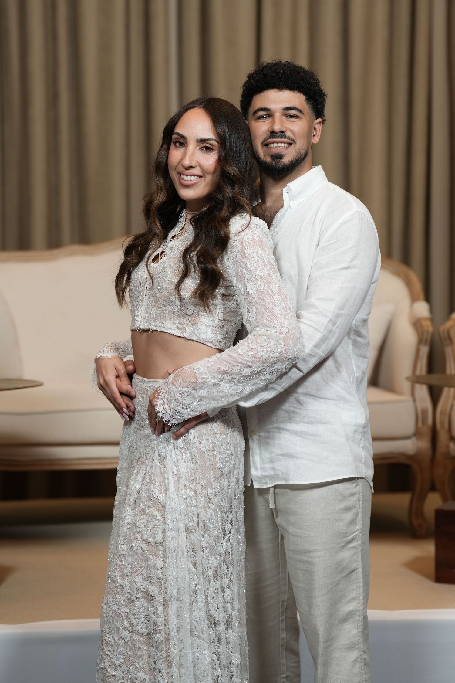 Couple in white outfits posing together, woman in lace lehenga and man in linen shirt, smiling in elegant interior.