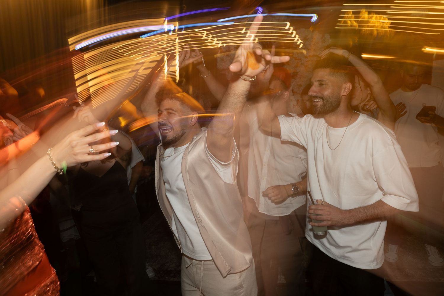 Group of young men dancing and celebrating at a nightclub party with dynamic light trails in background.