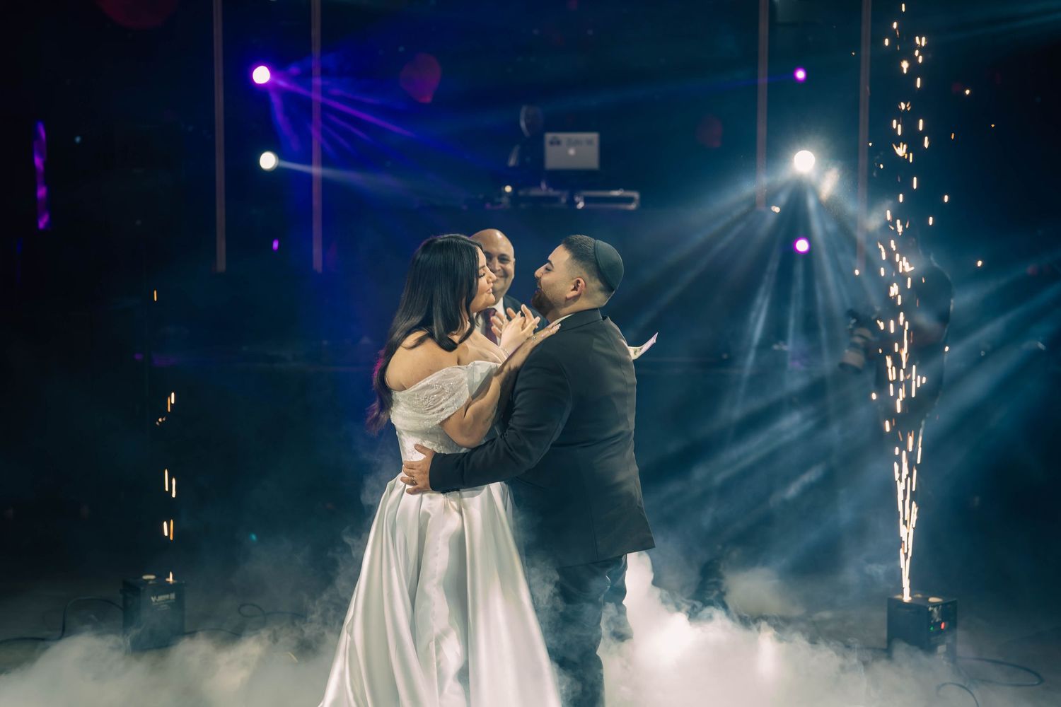 Couple shares romantic first dance surrounded by sparkling fireworks and dramatic fog on a dimly lit wedding reception dance floor.