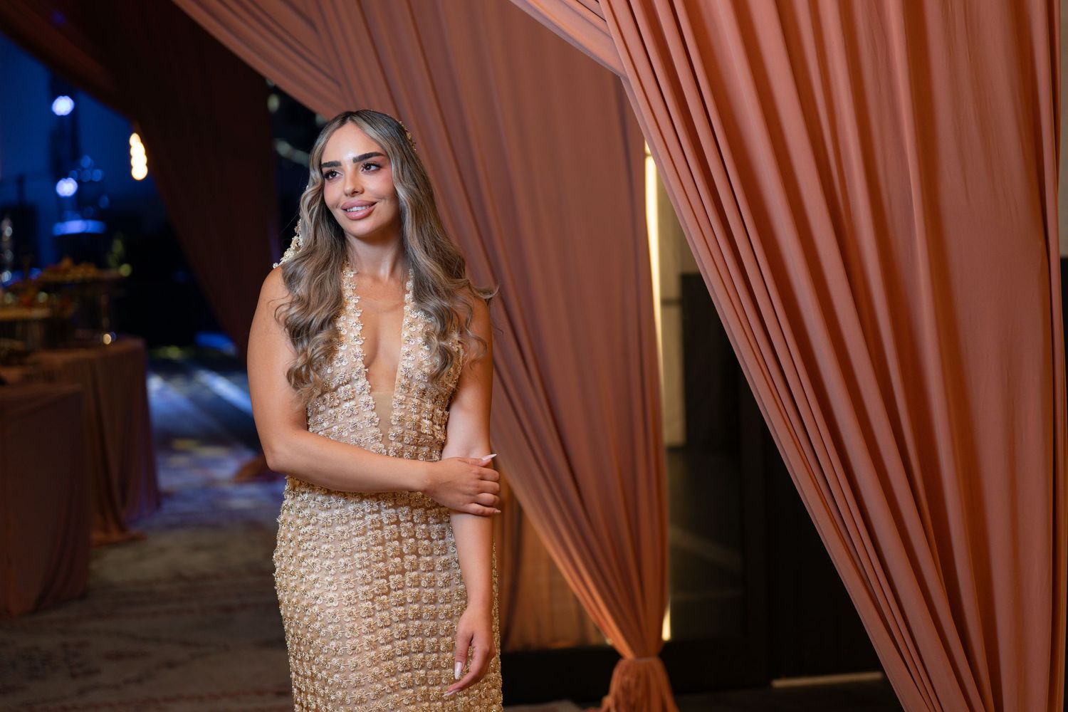 Woman in gold sequin gown poses elegantly by terracotta curtains at upscale evening event.