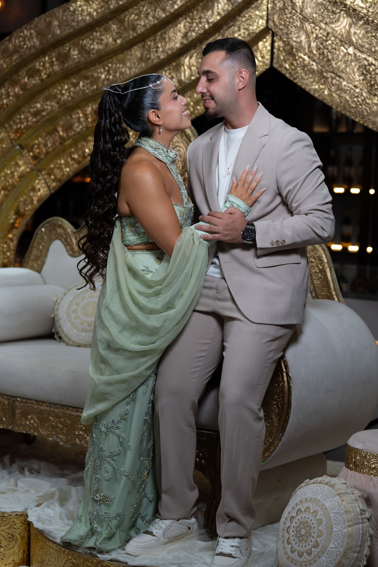 Couple in elegant attire embrace on ornate gold throne, woman in green sari, man in beige suit, at formal event.