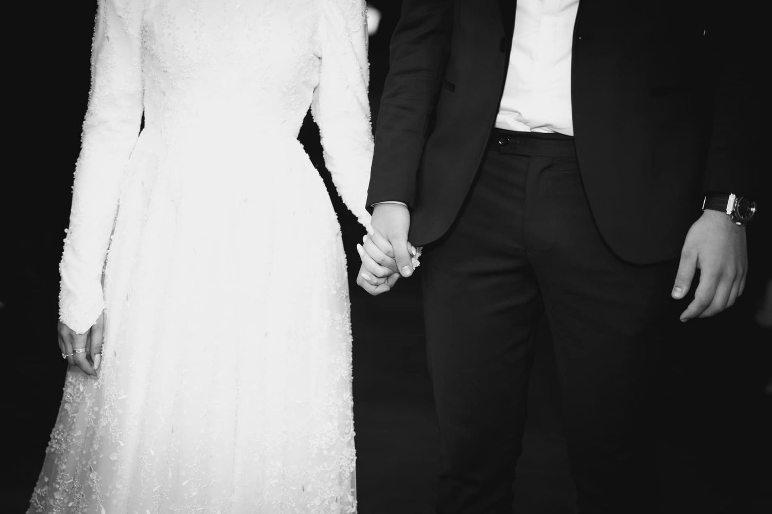Black and white photo showing couple holding hands in wedding attire.