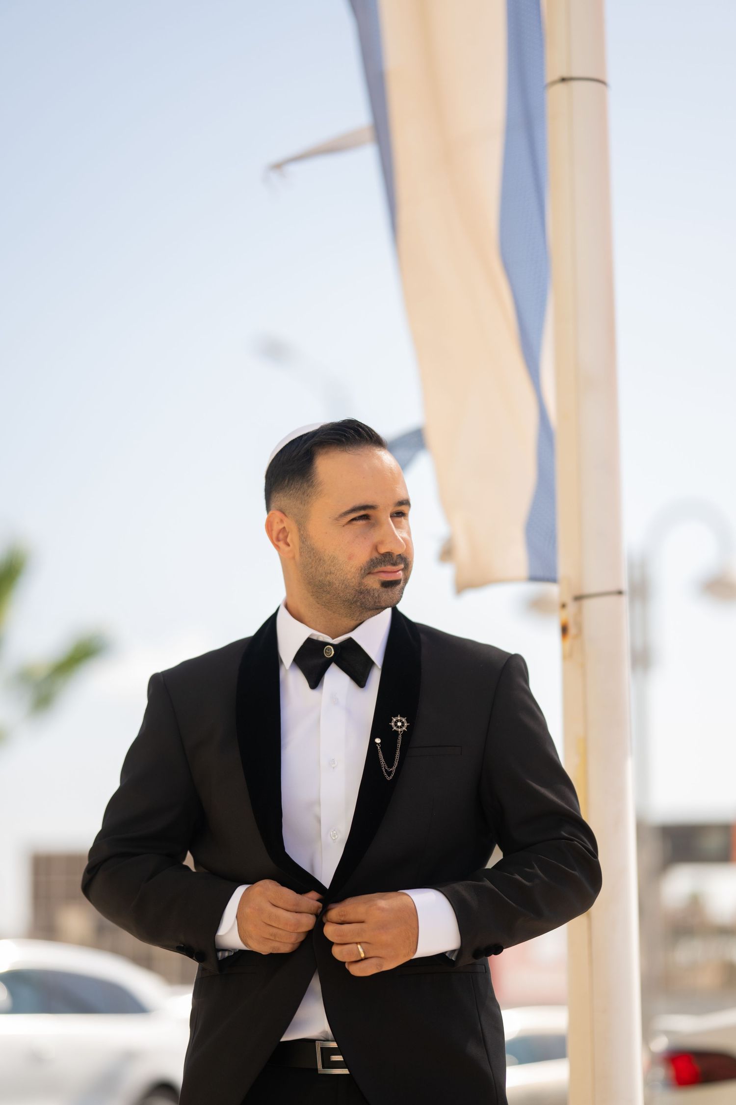 A groom in a classic black tuxedo adjusts his suit while standing outdoors under white fabric drapes.