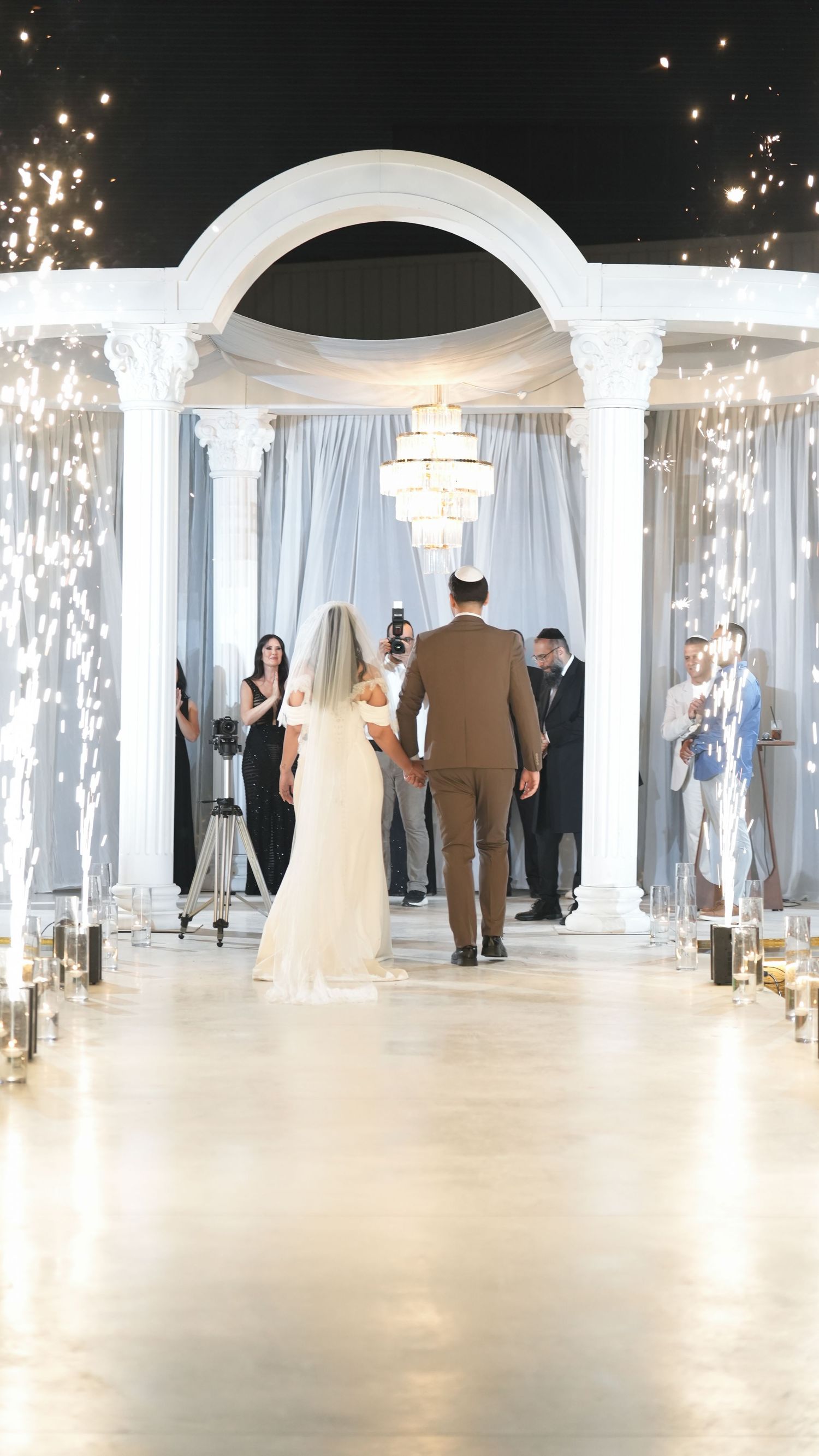 Bride and groom walk down aisle under white arch with sparkling fireworks at elegant wedding ceremony.