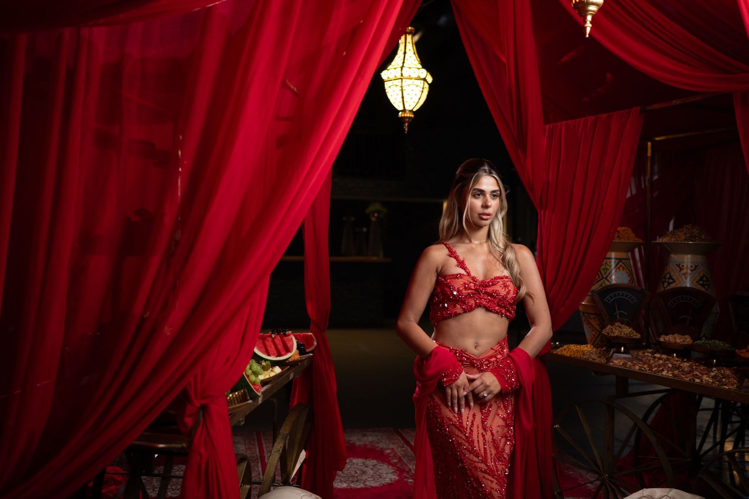 Woman in red belly dance costume poses between red curtains under warm lantern light in exotic setting.