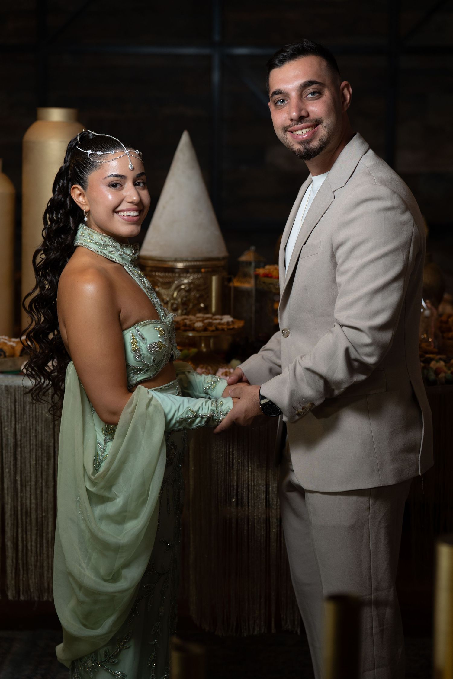 Elegant couple in formal attire holding hands, woman in green gown with headpiece, man in beige suit, at a decorated event.
