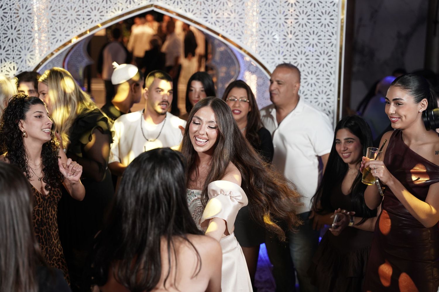 People dancing and socializing at a lively party or celebration with festive lighting in the background.