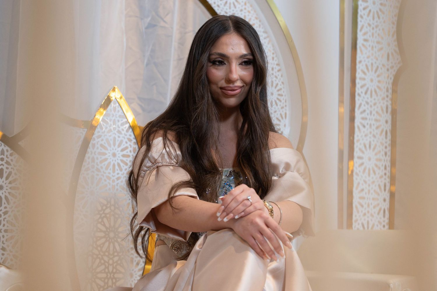 A woman in a glamorous cream-colored dress poses against an elegant beige backdrop with ornate wall details.