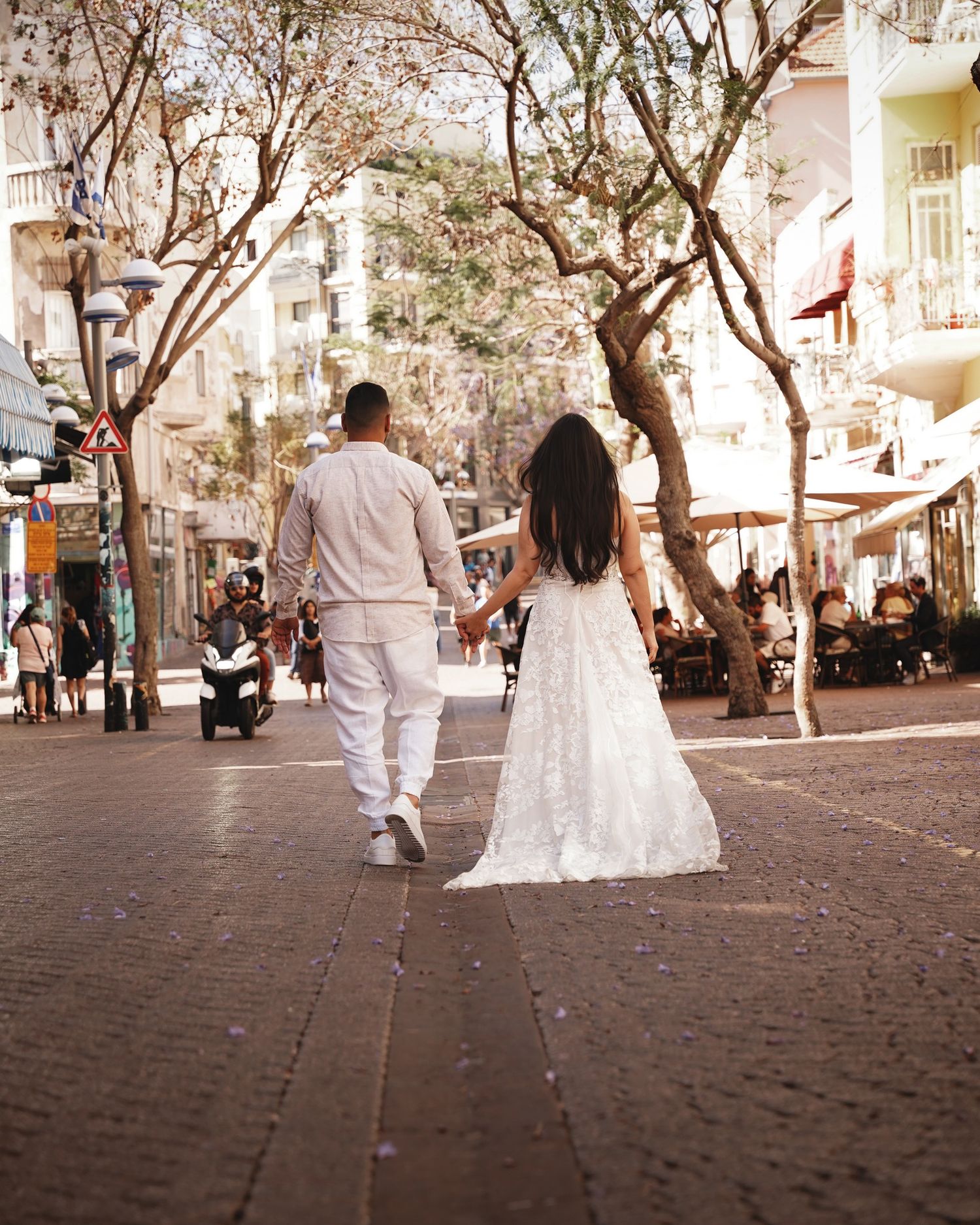 A couple in white attire walks down a tree-lined street on their wedding day.