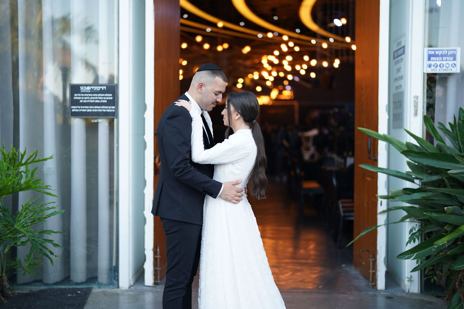 A romantic moment between newlyweds in front of a venue with twinkling lights.