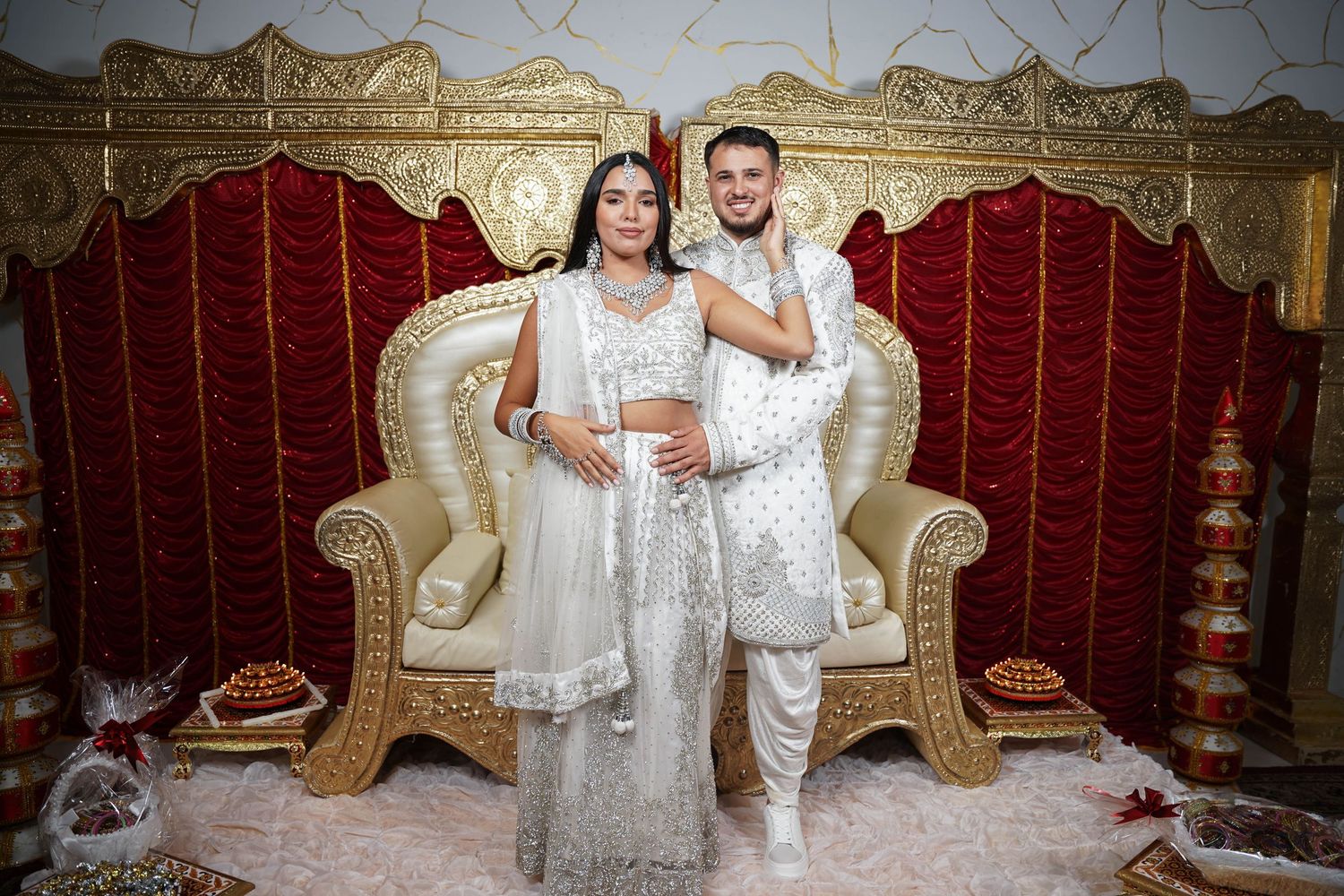 Wedding couple in matching white outfits seated on luxurious gold throne.