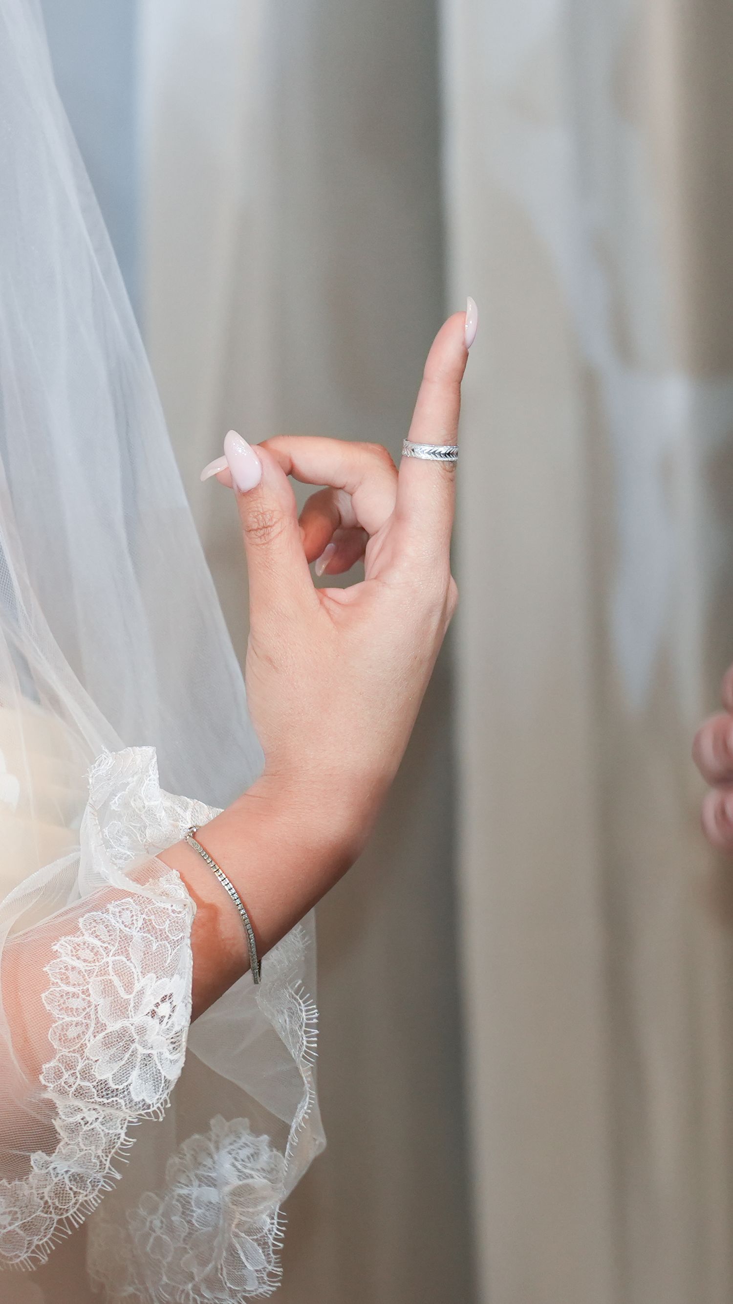Bride's hand holding a white lace veil, adorned with silver rings and a diamond bracelet, with manicured pink nails.