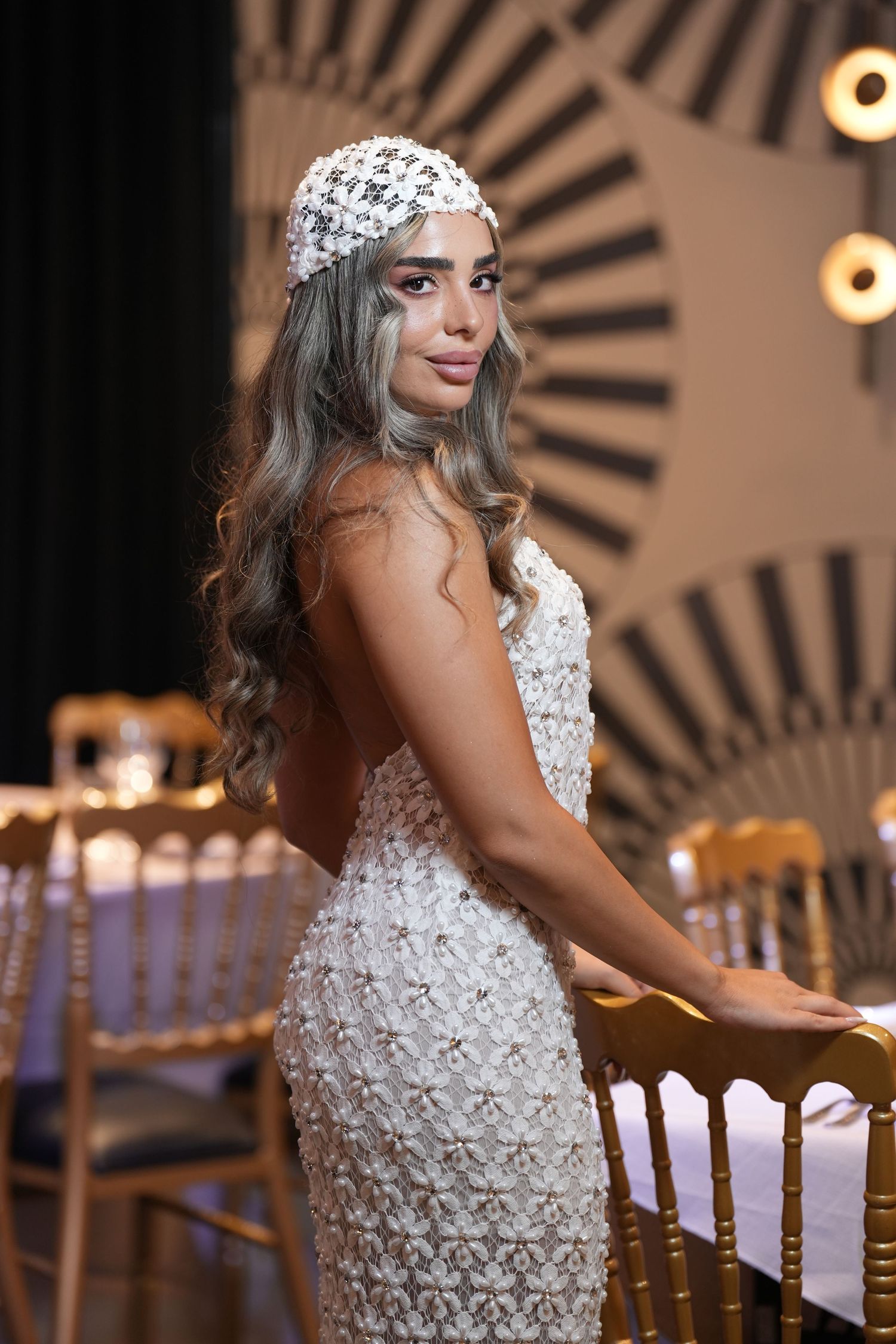 Elegant woman in white floral gown and pearl headpiece poses gracefully at a formal event venue.