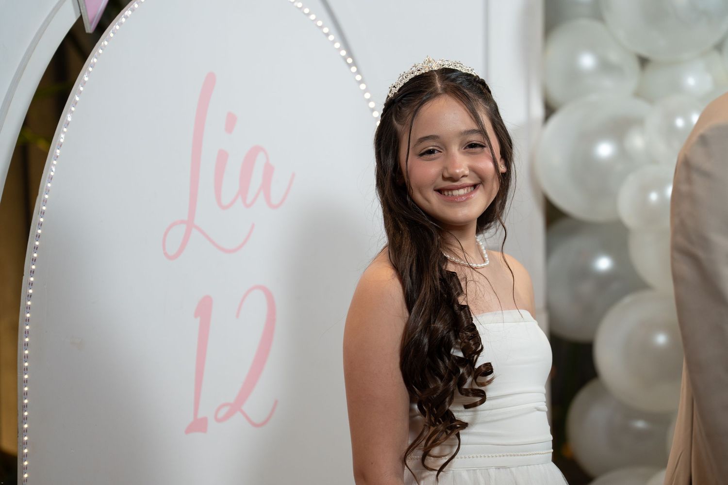 A young person in a white dress poses in front of white balloons during what appears to be a quinceañera celebration.