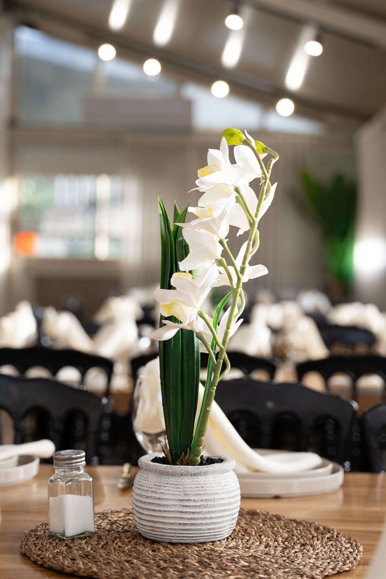 White lilies in modern ceramic vase centerpiece on woven placemat with dining setup.