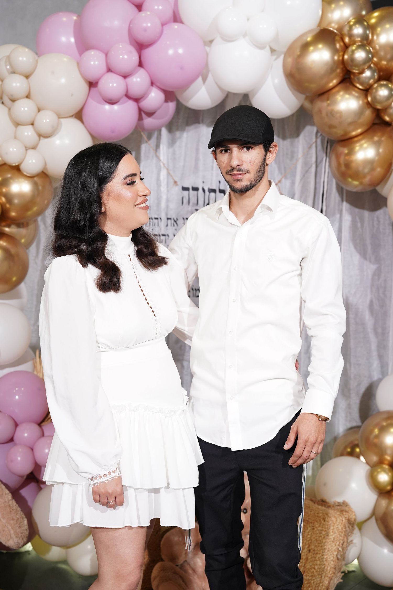 Couple in matching white outfits posing against balloon arch backdrop.