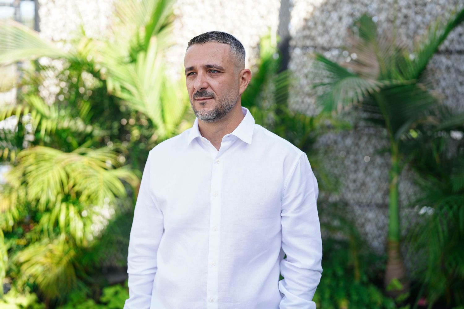 Man in crisp white shirt stands in front of tropical garden landscape.
