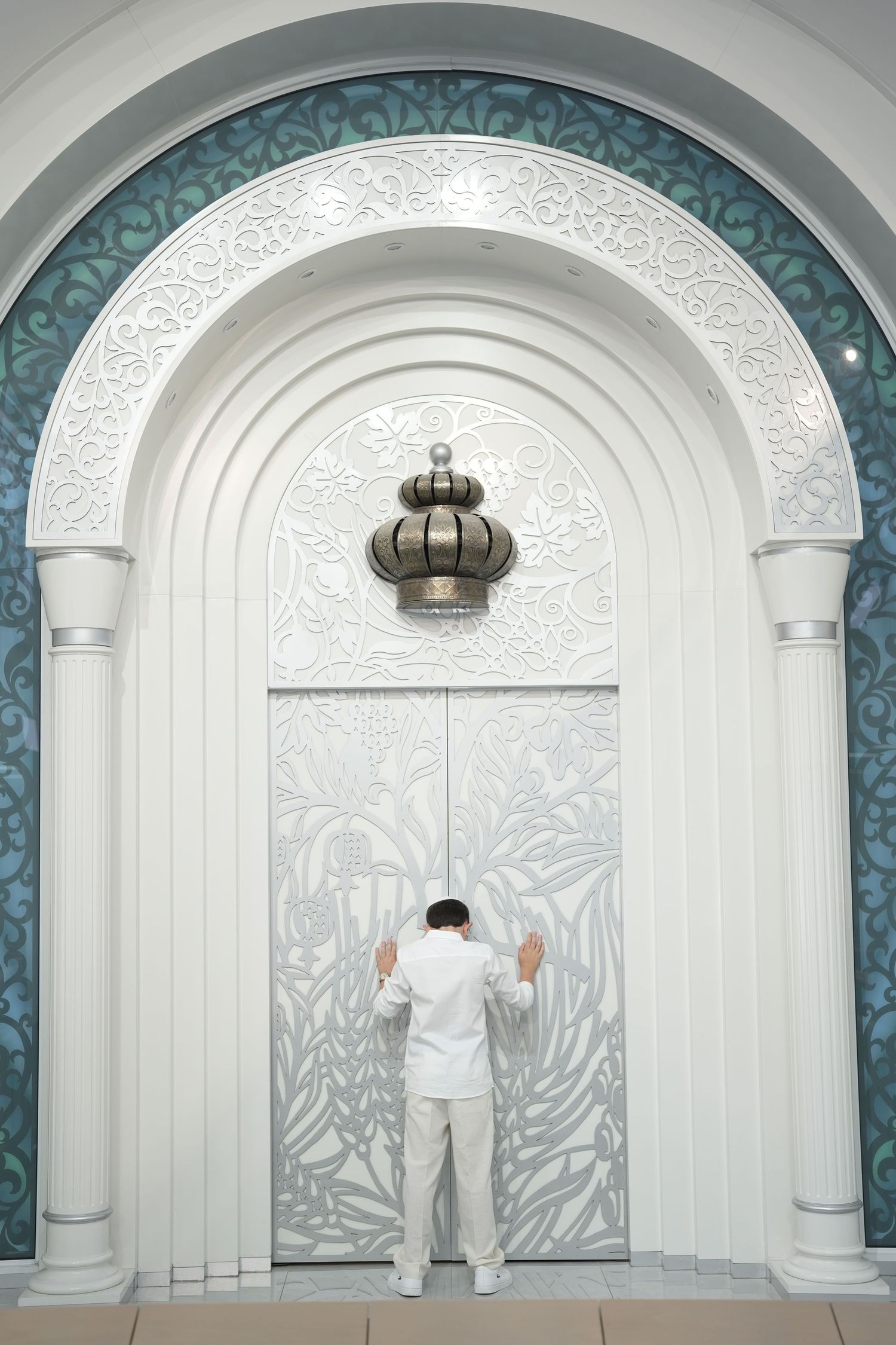 Man in white attire standing before ornate arched mosque door with decorative silver panels and lantern.
