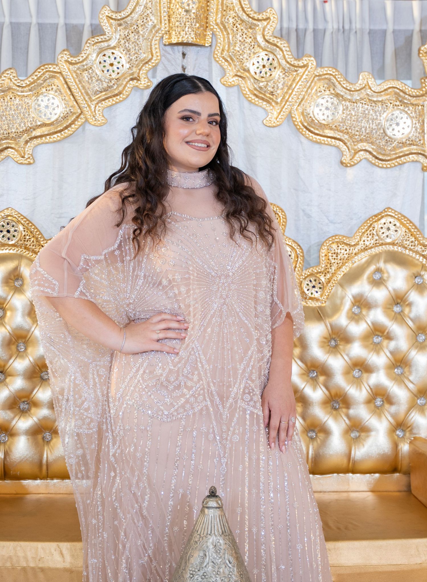 Full-length shot of person in flowing blush evening gown against elegant gold throne setting.