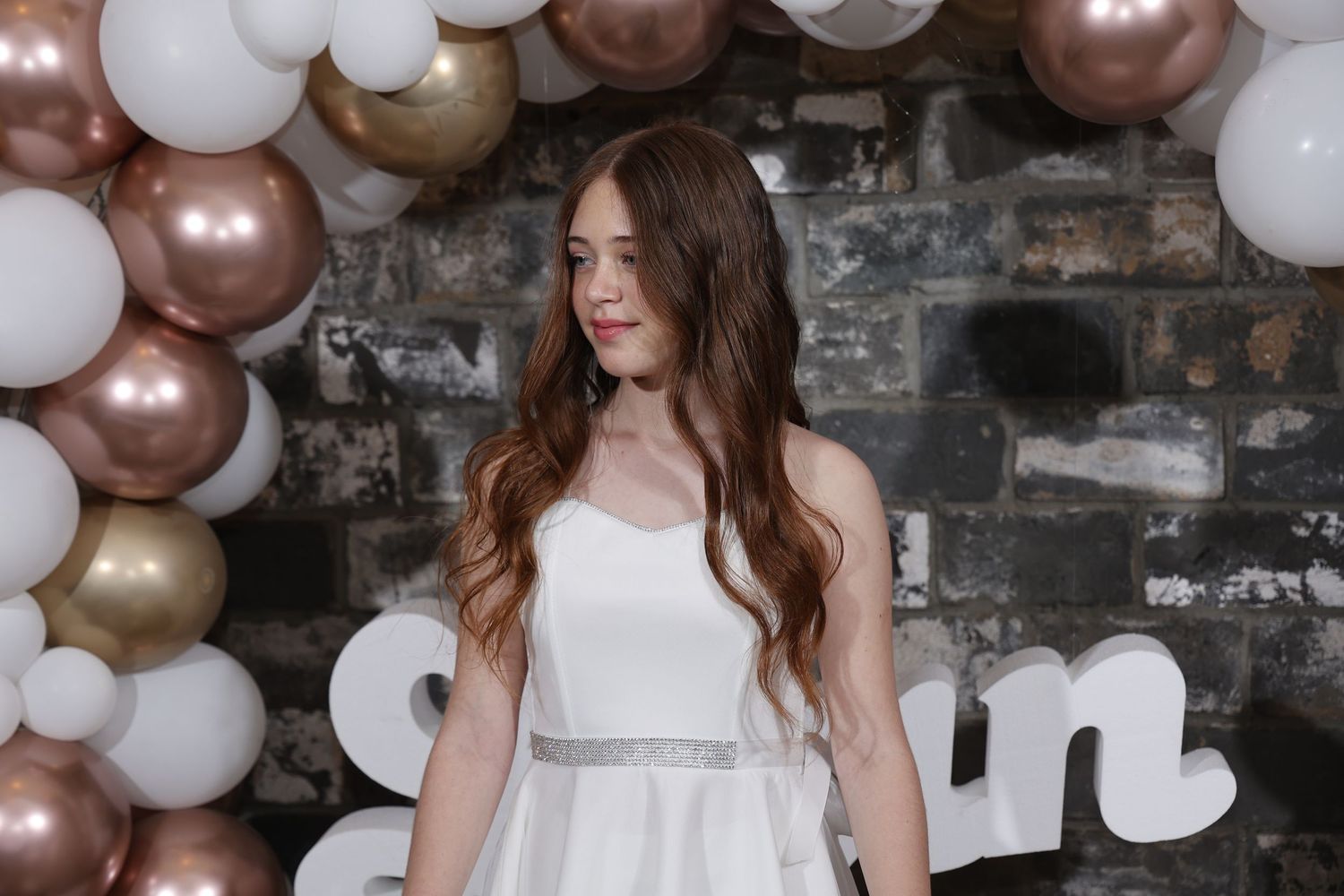 Elegant party setup with rose gold and white balloon arch against brick wall.