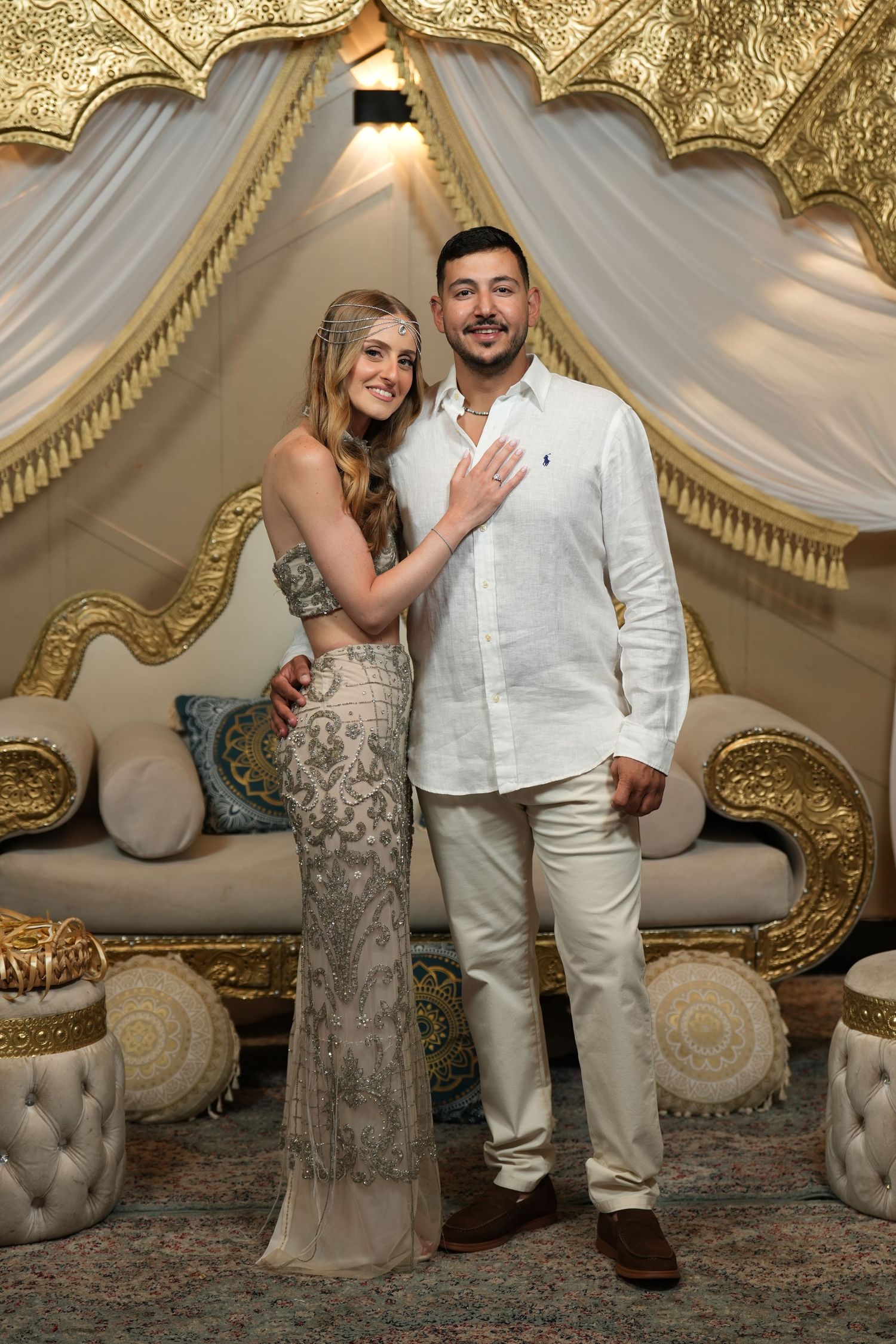Couple posing elegantly in ornate gold and white decor, woman in beaded gown, man in white linen shirt and pants.