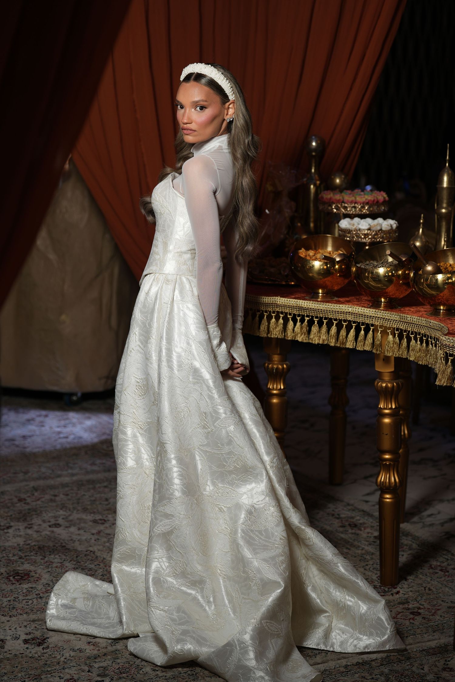 Elegant bride in a white long-sleeve gown with veil headpiece stands near a ornate golden table with candles.