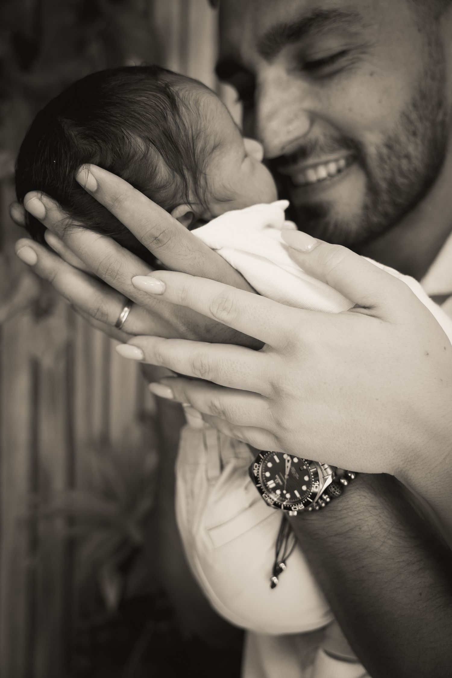 Black and white photo of a smiling father tenderly holding his newborn baby, wearing a wedding ring and watch.