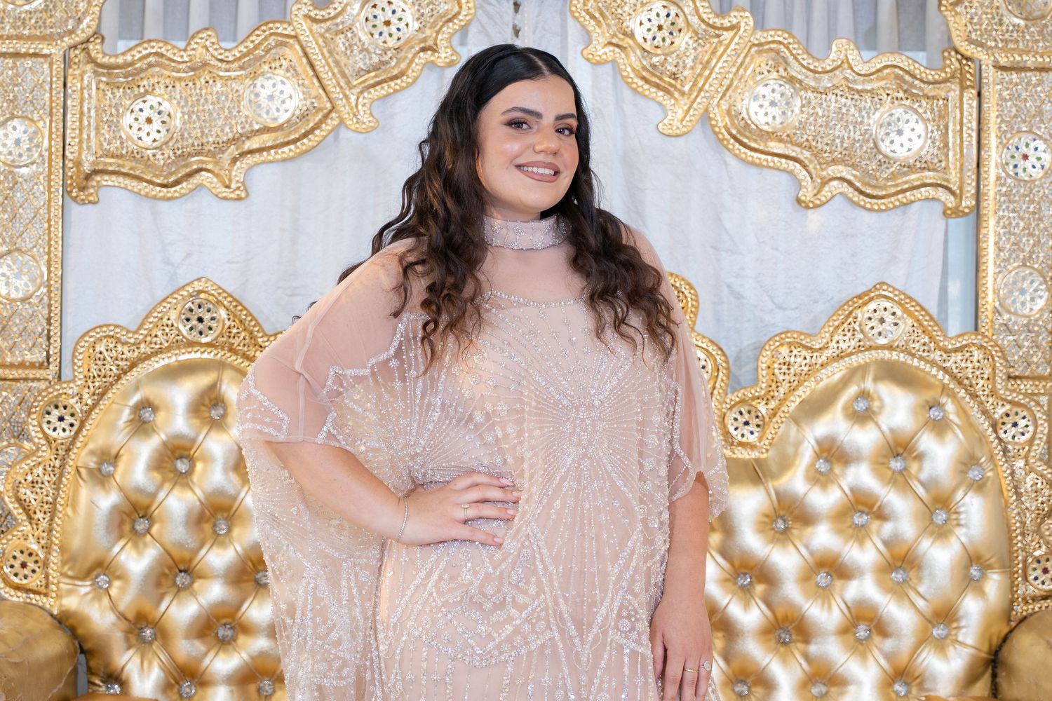 Person poses in shimmering pink dress against ornate gold and white backdrop.