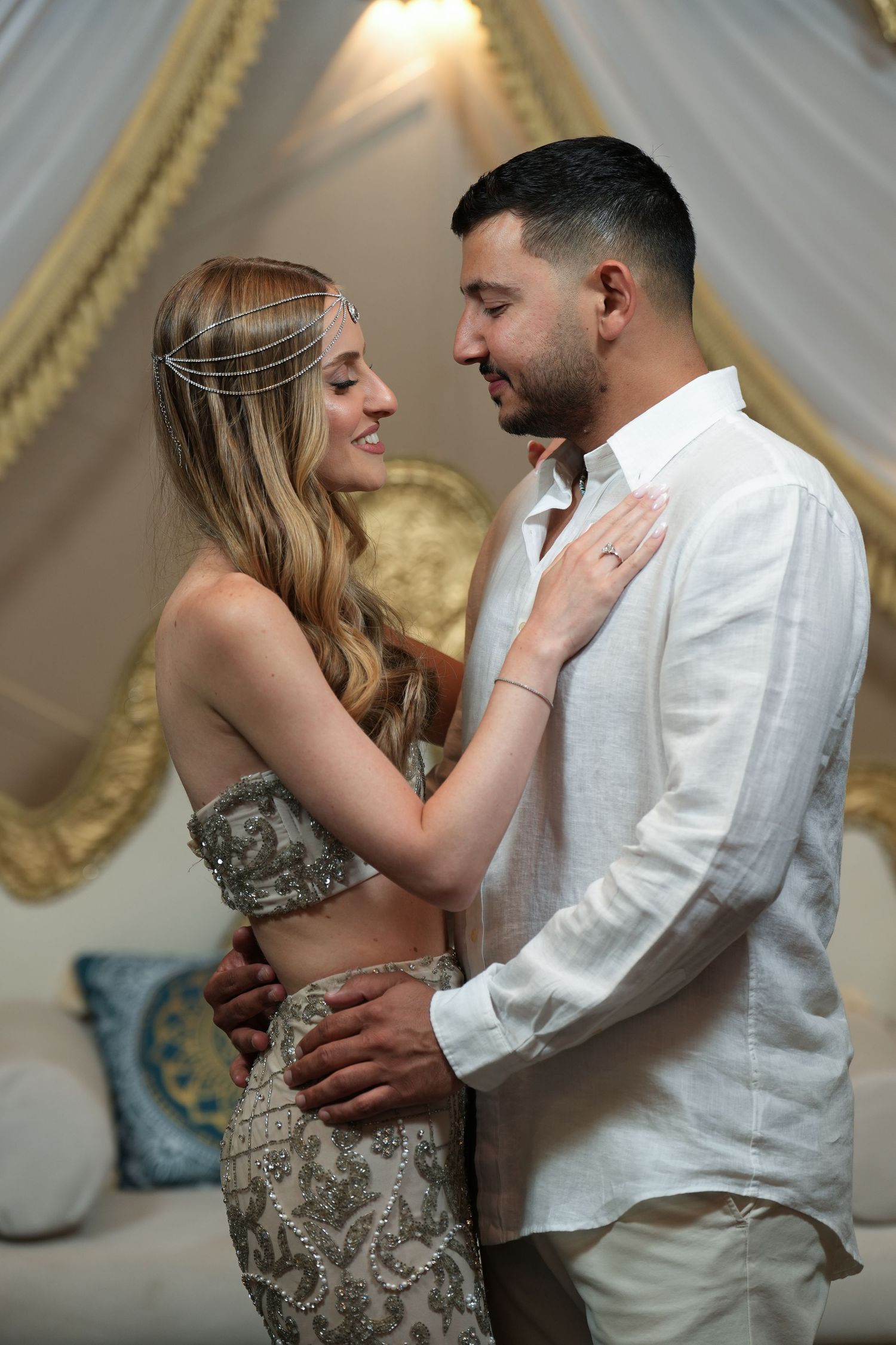 Couple in elegant attire sharing a romantic moment, woman in embellished crop top with head chain, man in white linen shirt.
