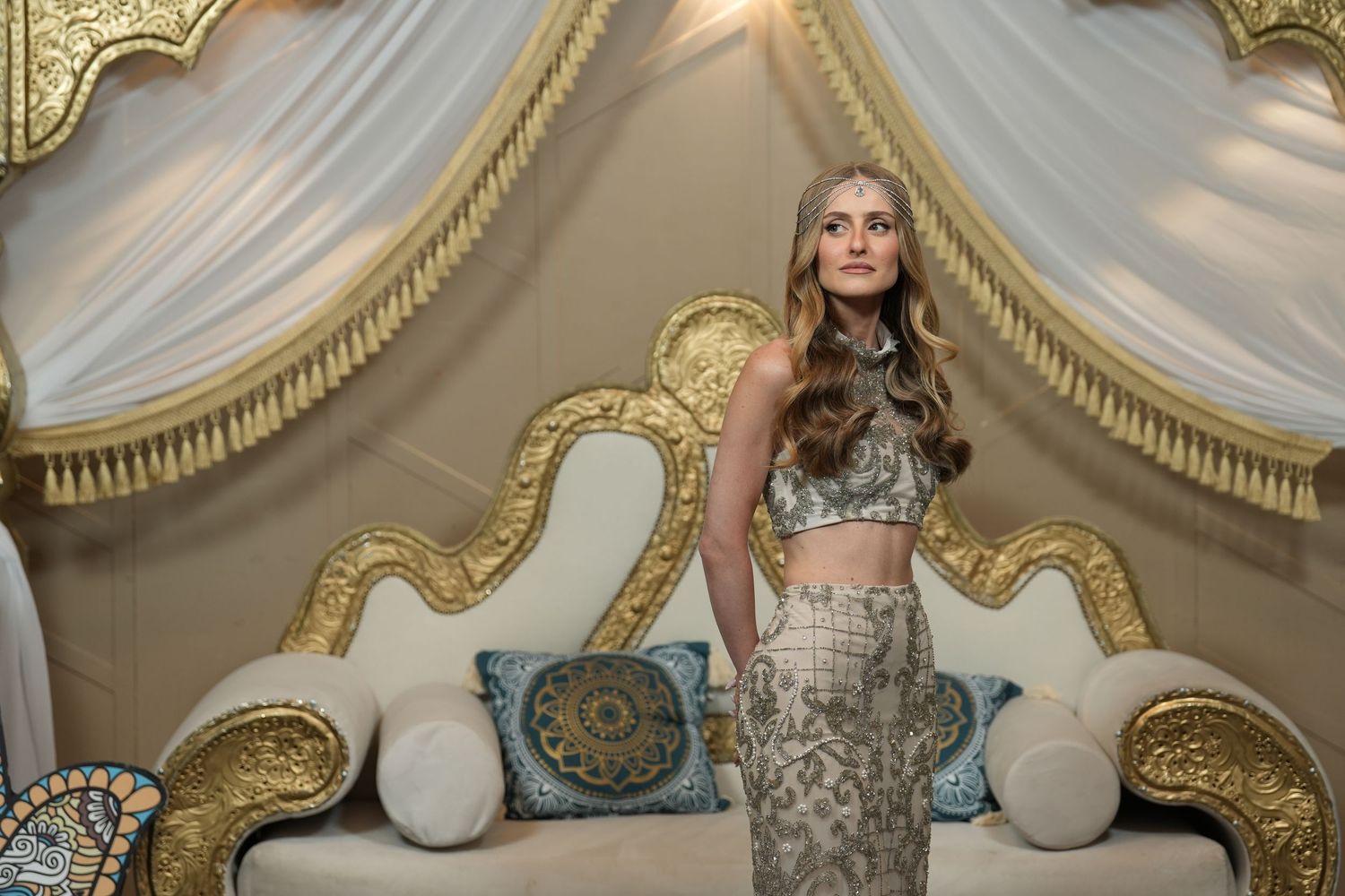 Woman in elegant silver crop top and skirt poses before ornate gold-framed sofa with decorative pillows.