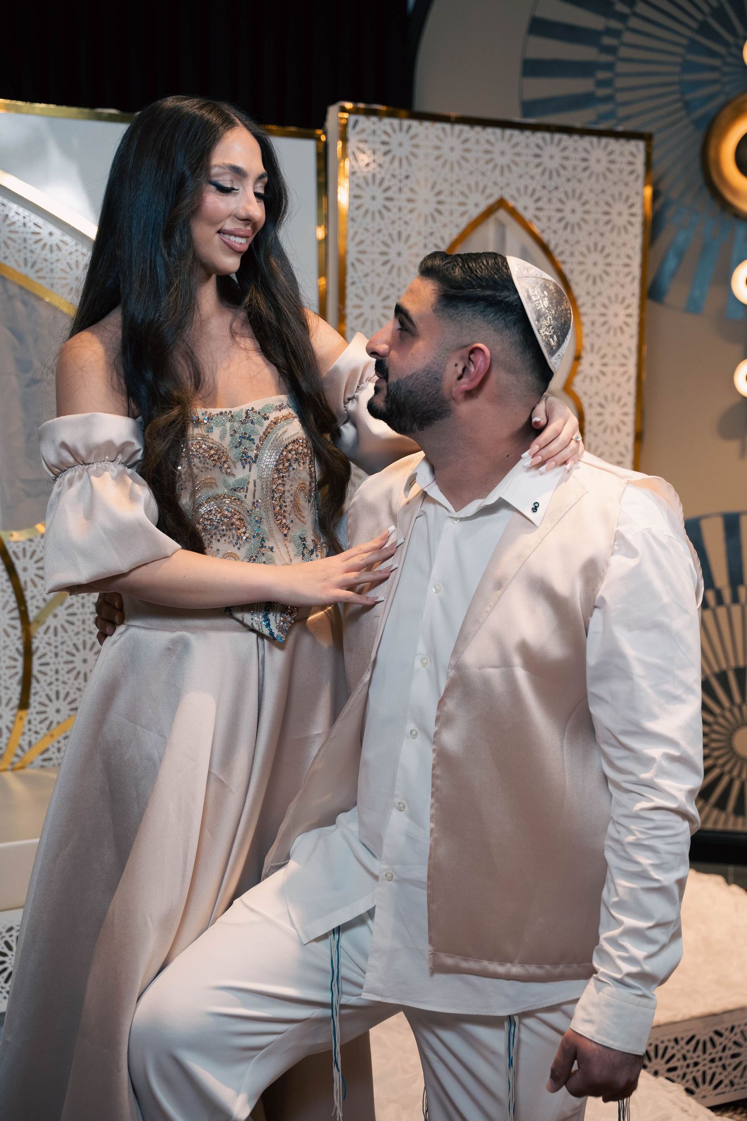 A couple in elegant white and champagne-colored outfits share an intimate moment at a formal event.