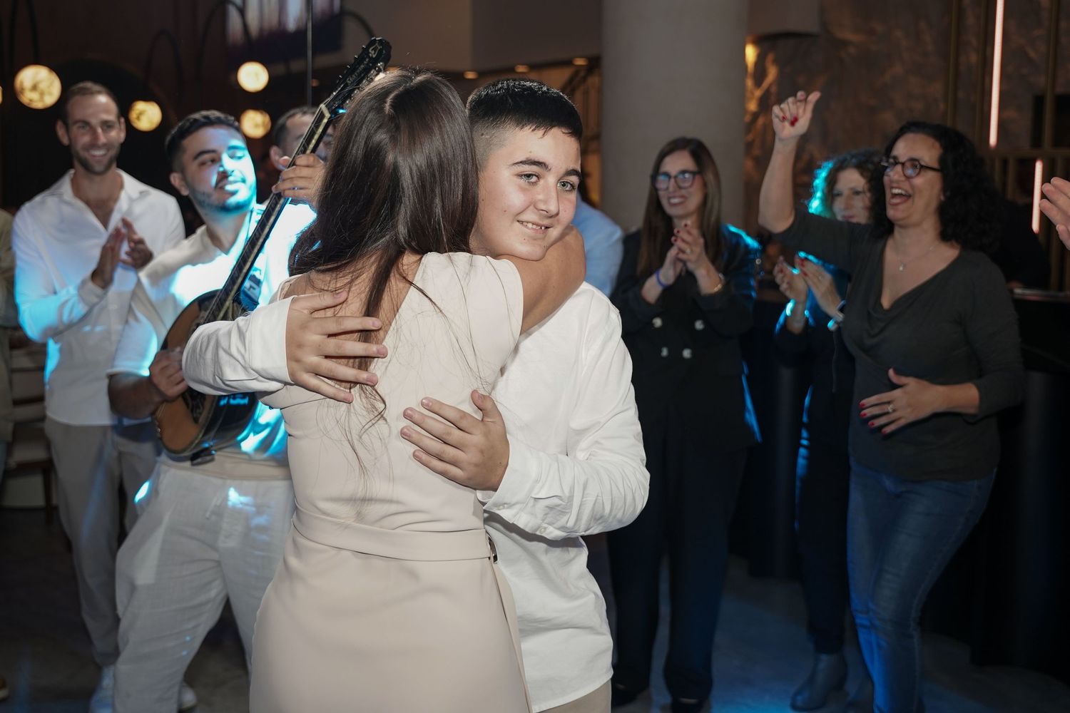 Two people embrace in a warm hug while party guests look on and applaud during a celebration event.