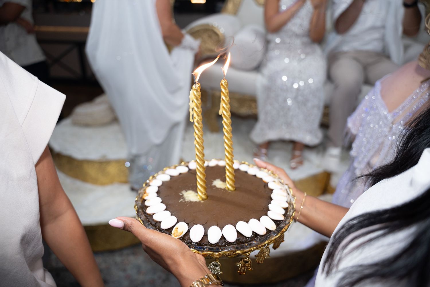 Hands holding a chocolate birthday cake with two lit gold candles, surrounded by people in white outfits.