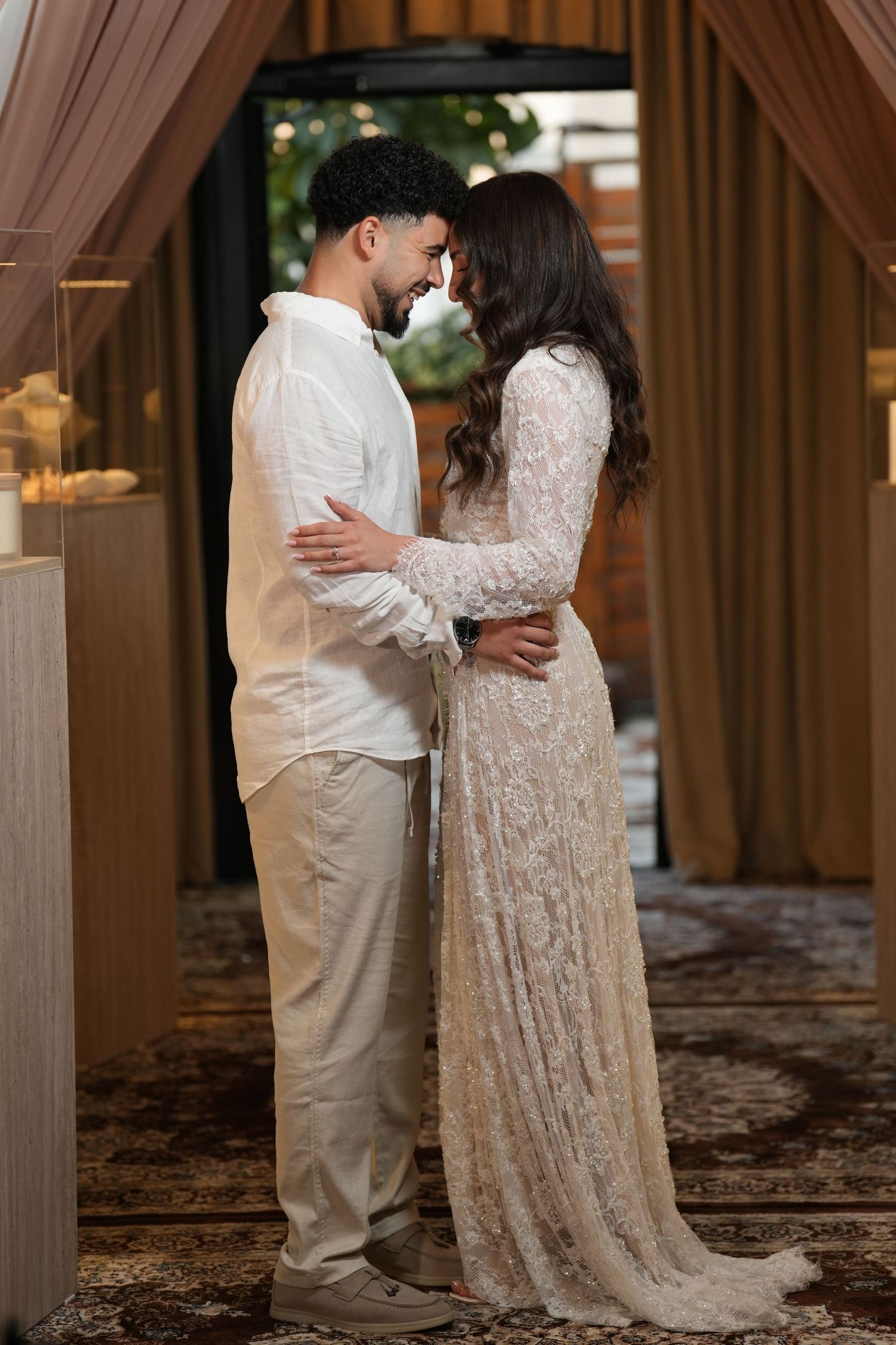 A couple in elegant white wedding attire shares an intimate moment, standing face to face in a warmly lit venue.