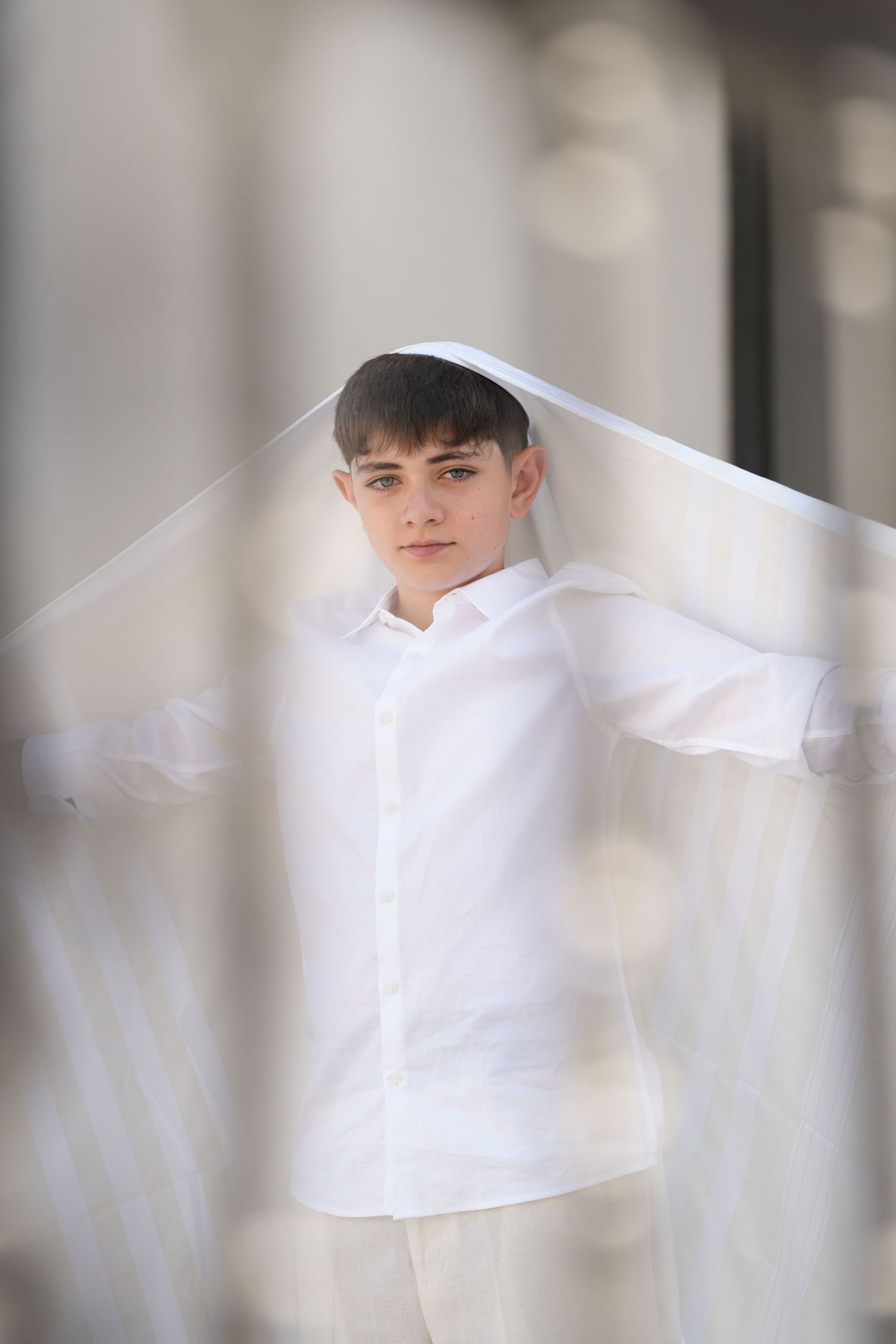 Young boy in white shirt holds sheer fabric overhead outdoors in elegant portrait photography.