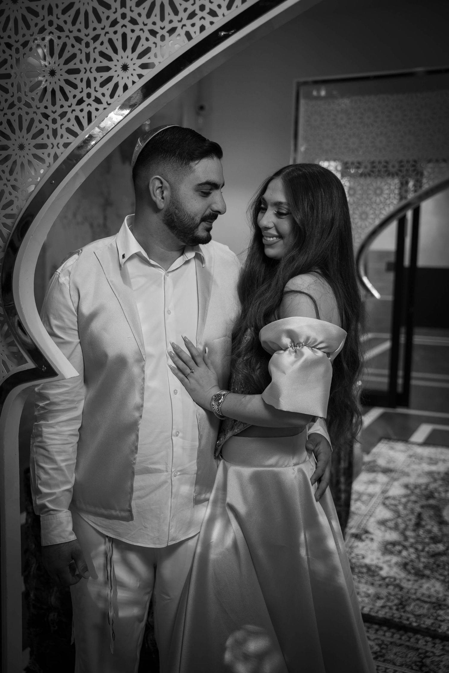 A black and white romantic photo of a couple in elegant attire sharing an intimate moment under an ornate archway.