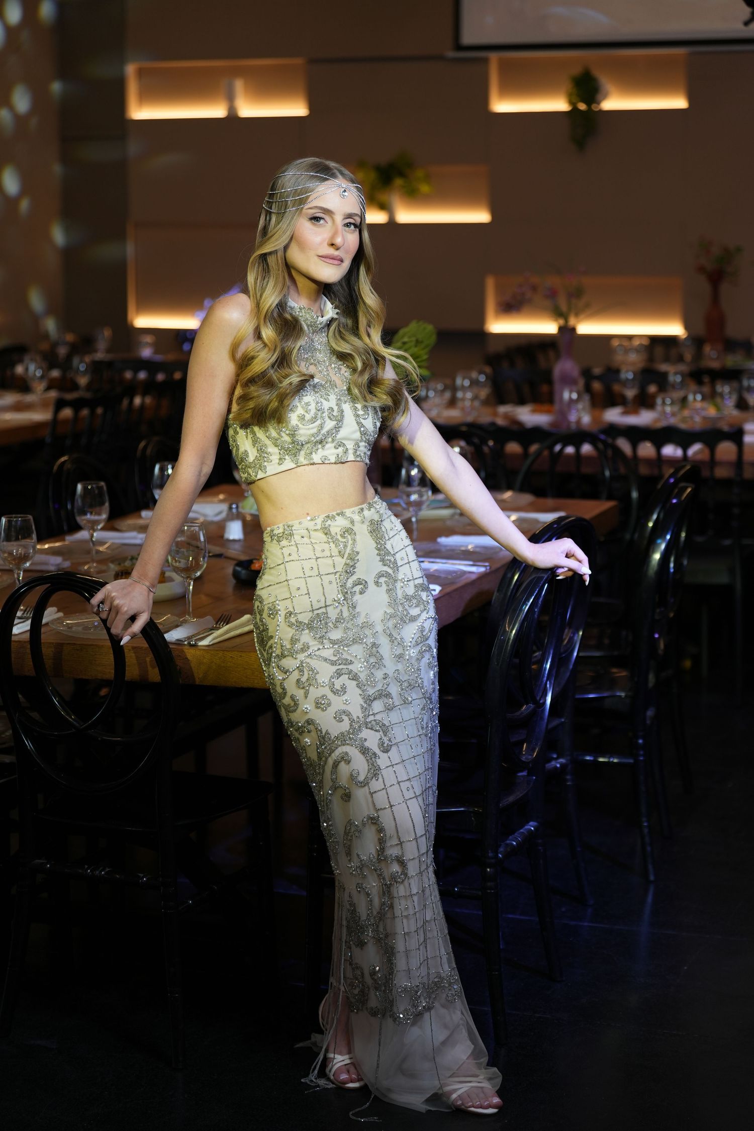 Young woman in elegant gold beaded two-piece gown posing in a dimly lit upscale restaurant setting.