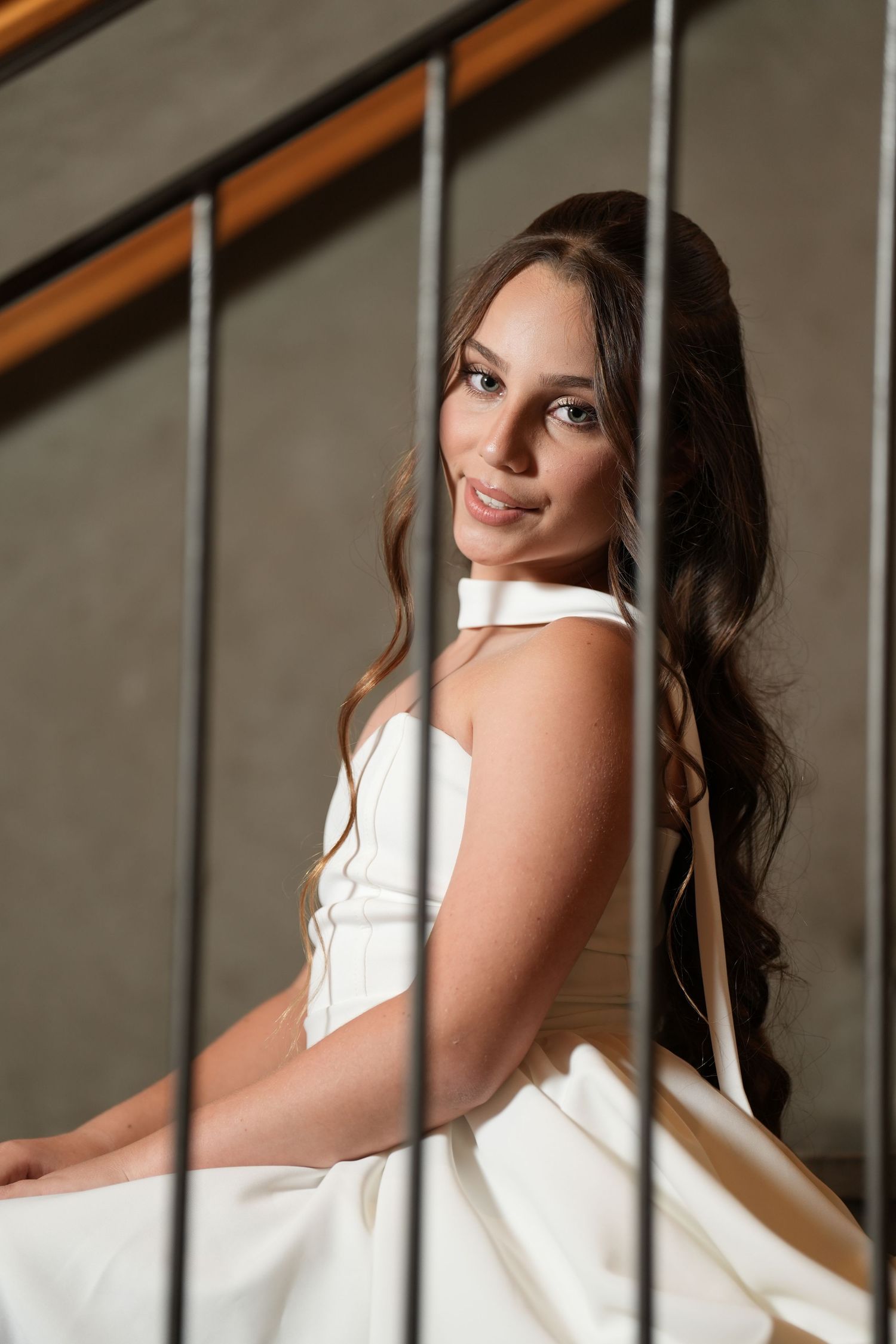 A bride in a white gown sits gracefully on stairs, smiling softly with long wavy brunette hair.