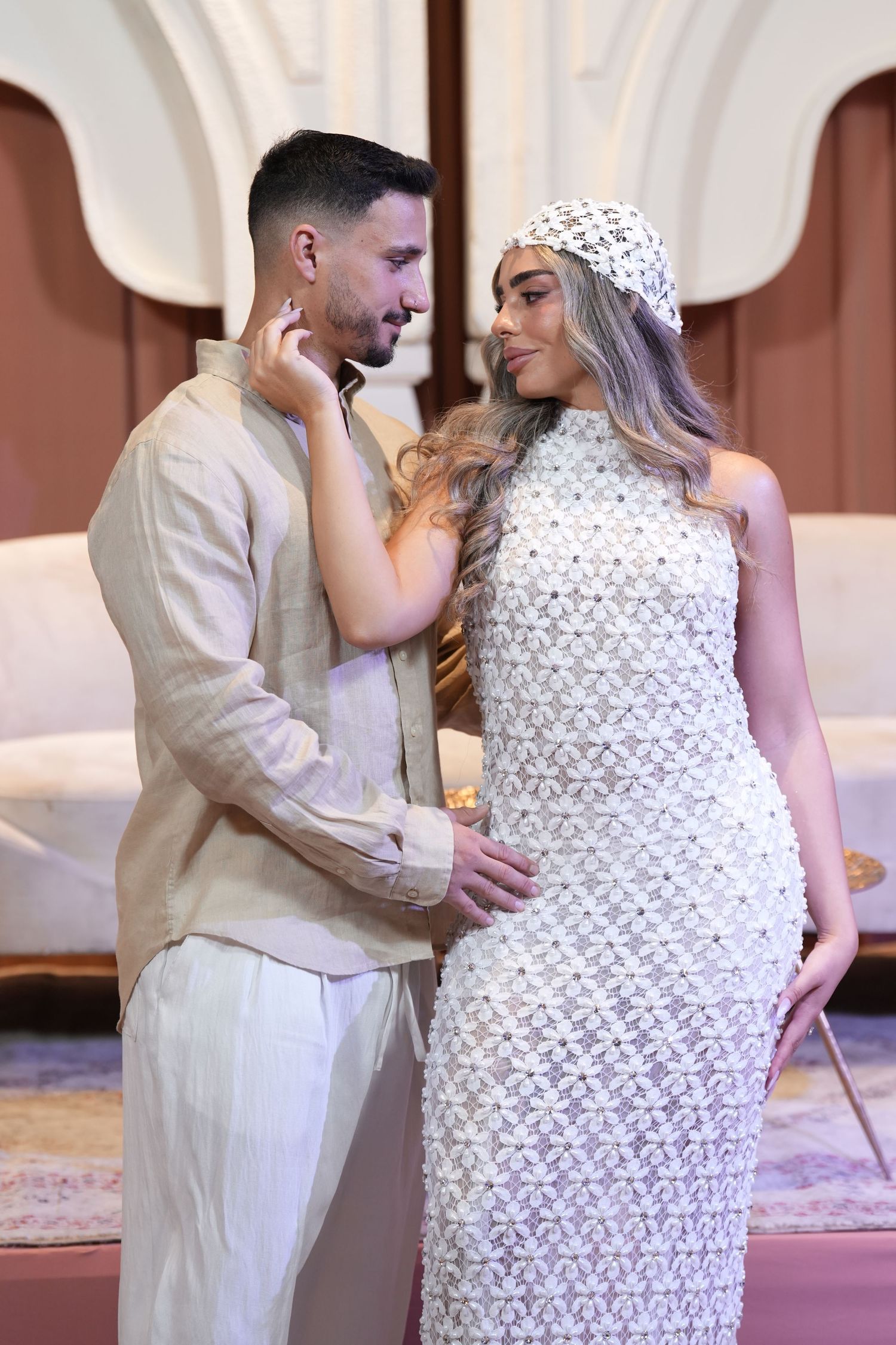 A couple in elegant attire share a romantic moment, woman in white textured dress and hat, man in beige outfit.