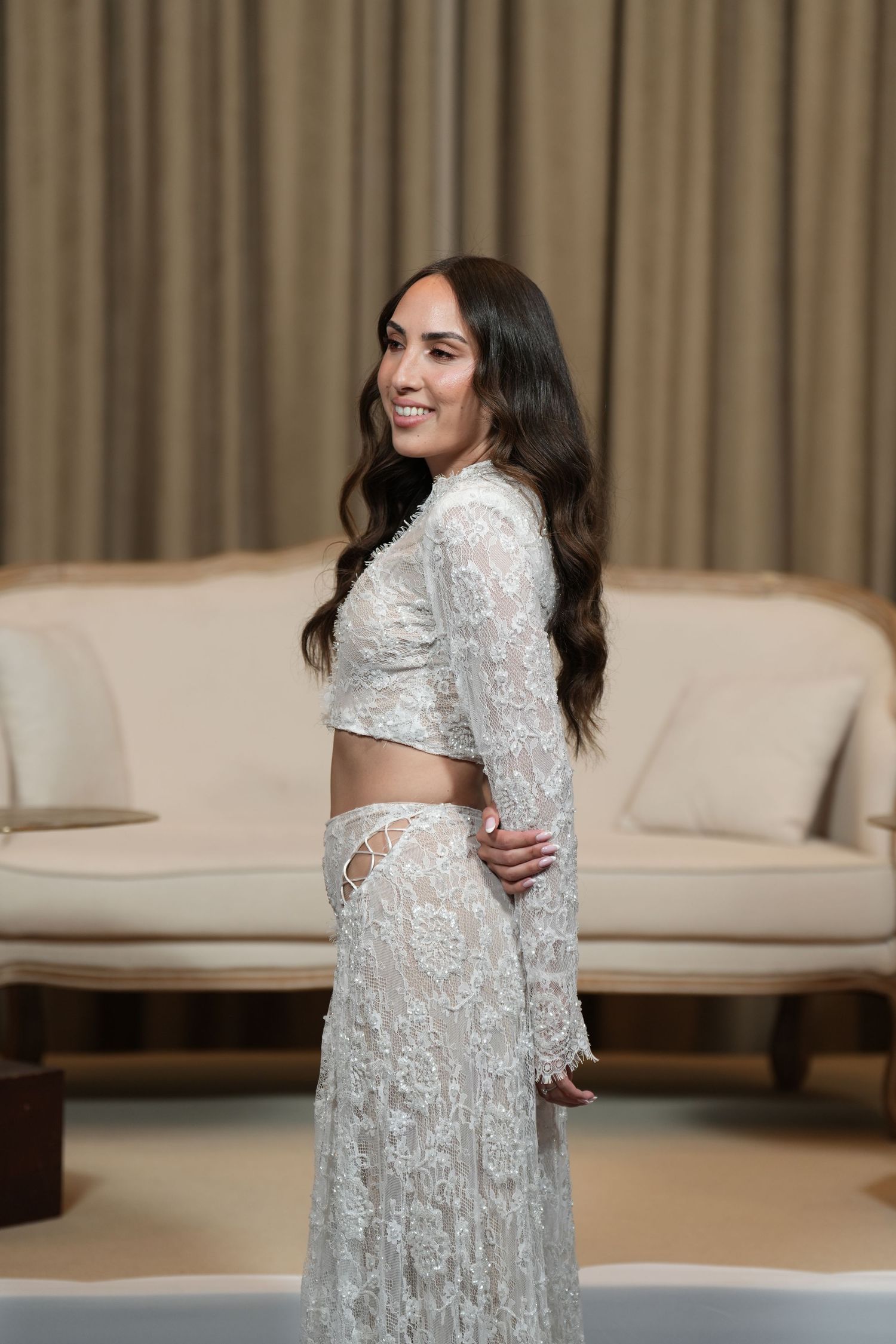 Woman in white lace two-piece outfit posing elegantly in a luxurious cream-toned room.
