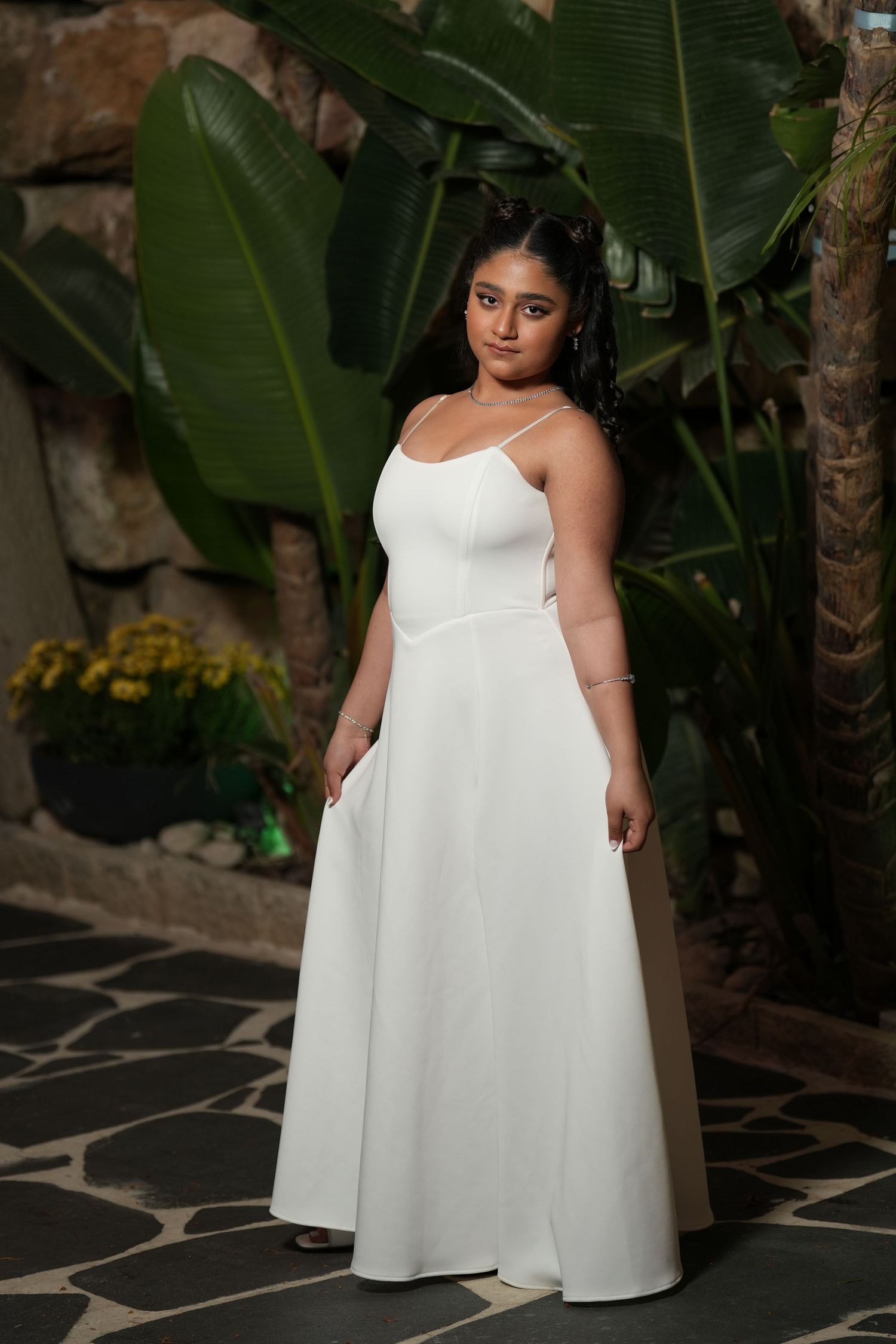 Woman in elegant white spaghetti strap A-line gown posing in front of tropical plants and stone wall.