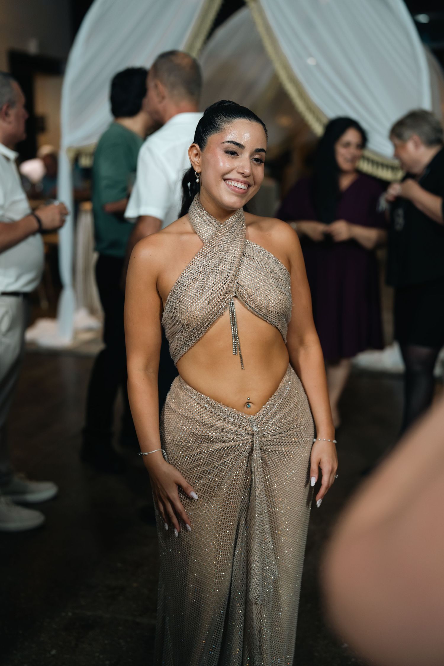 A smiling woman in a glamorous gold halter dress poses at an elegant event with guests in the background.