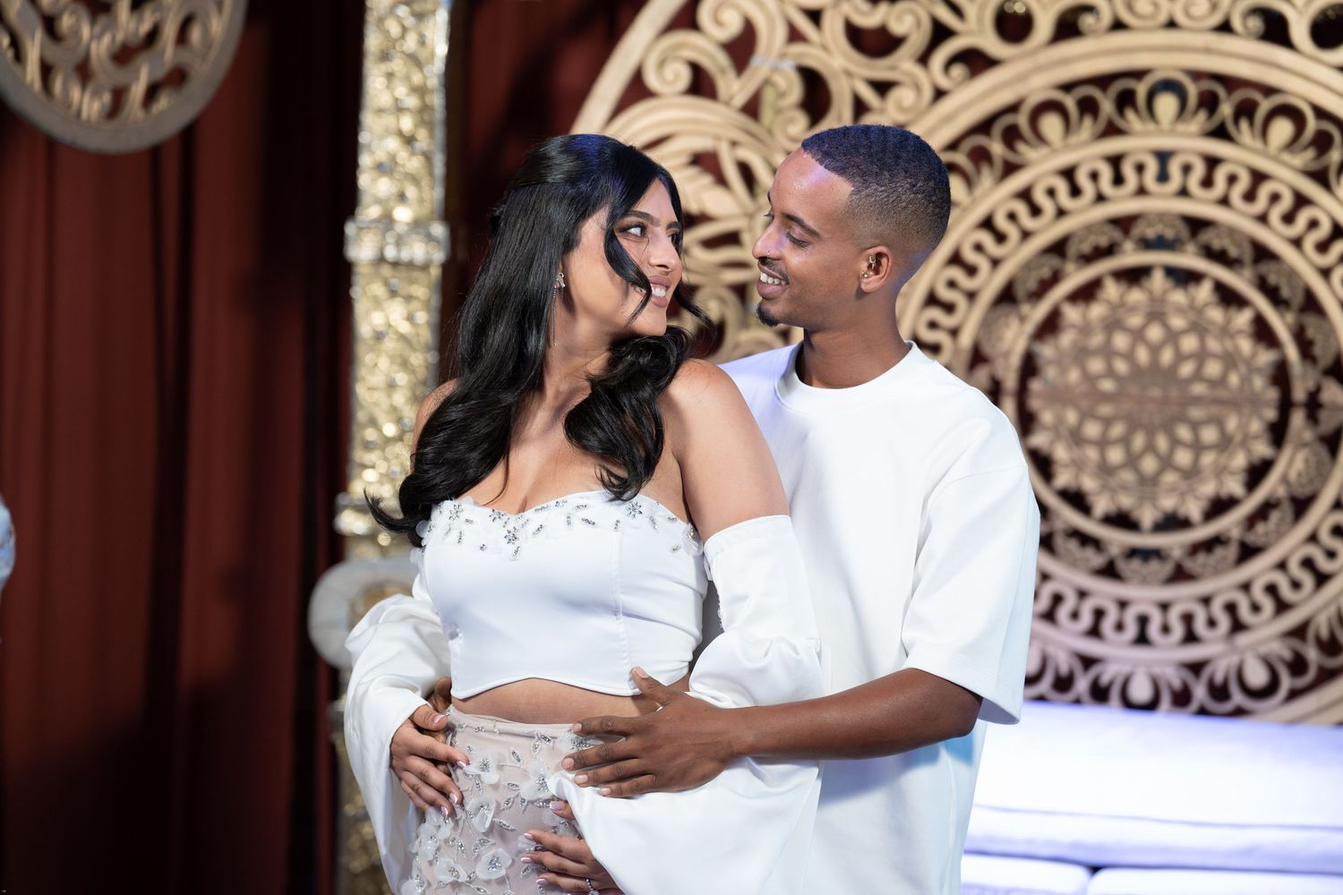 Couple in matching white outfits posing together in front of ornate gold decor backdrop.