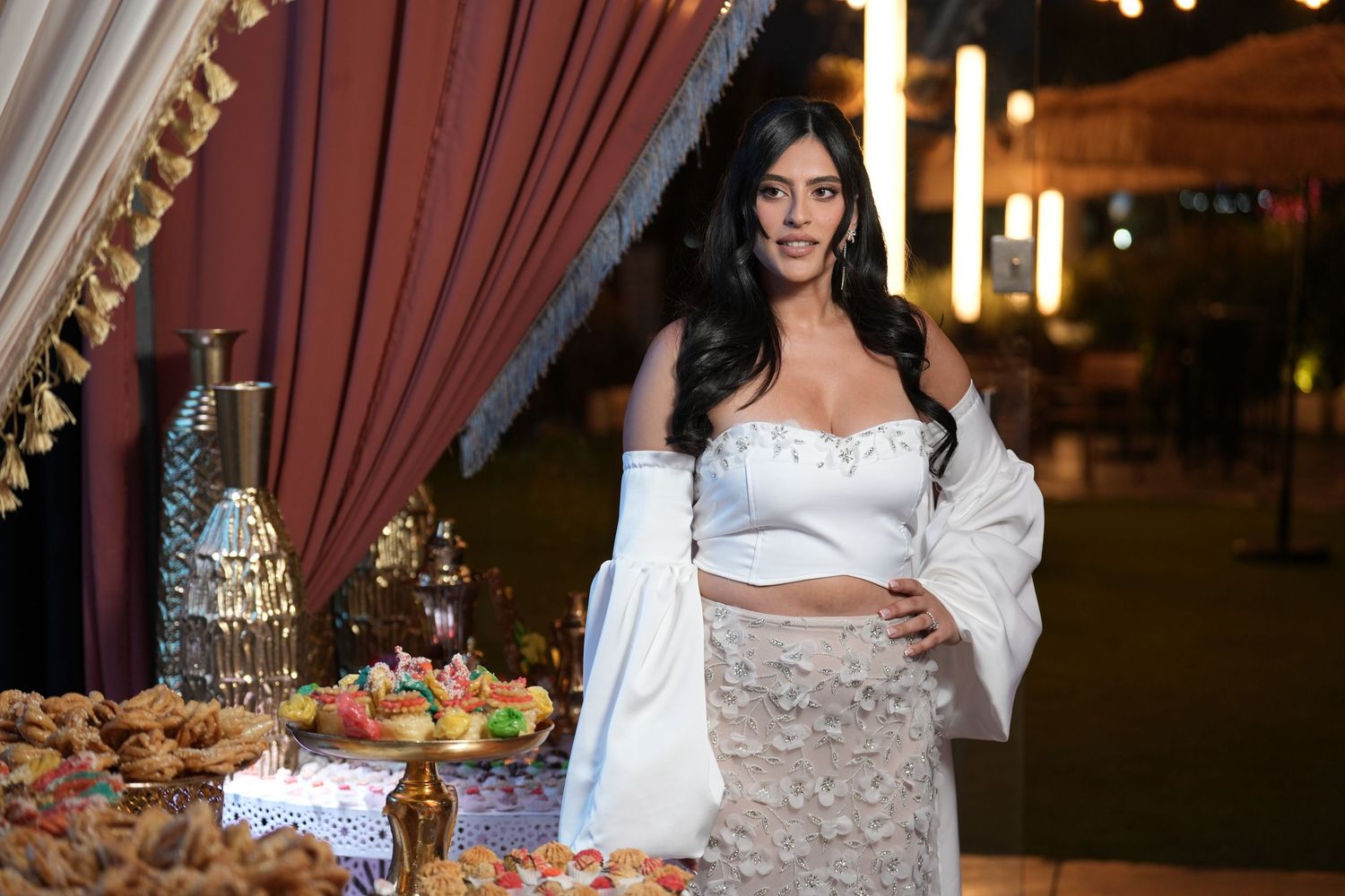 Woman in white crop top and skirt poses elegantly beside a festive dessert table with colorful treats and decorations.