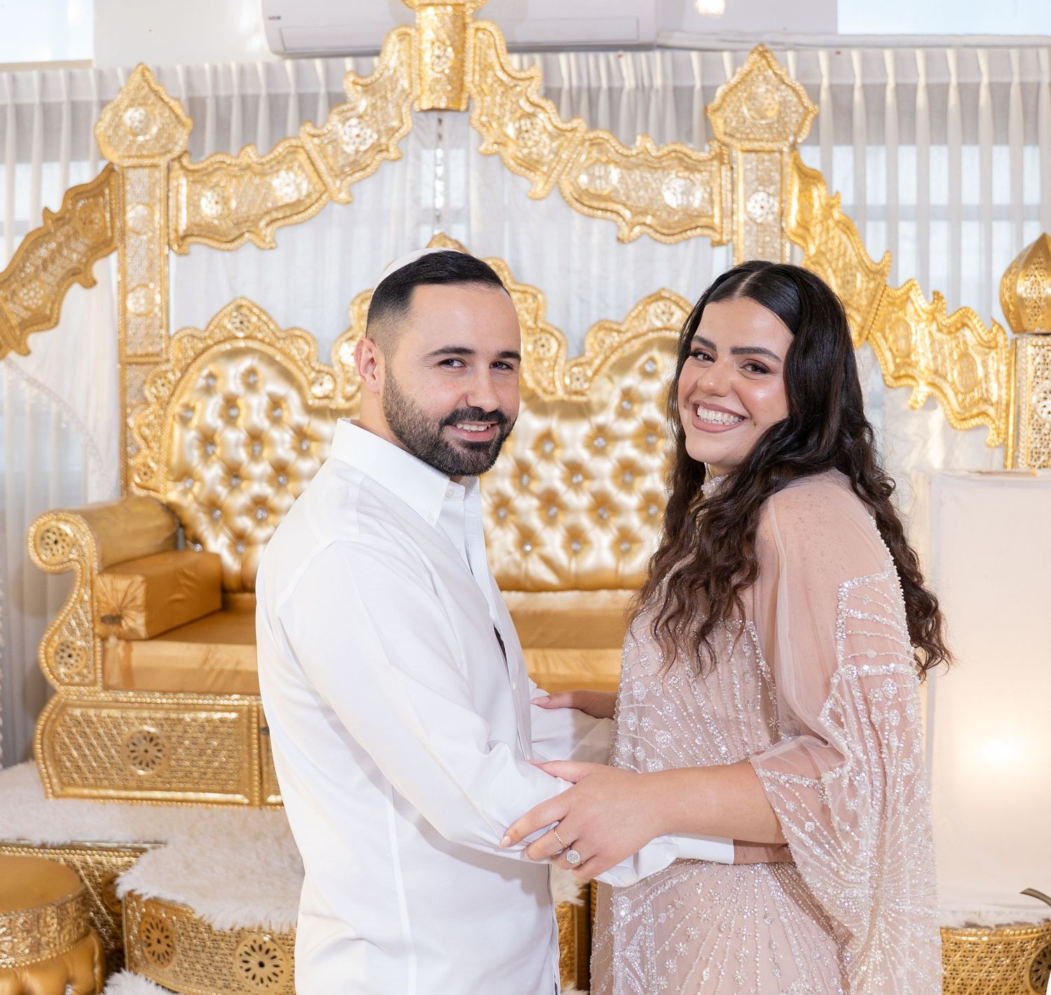 Romantic couple poses in front of ornate gold backdrop wearing elegant white and blush formal attire.