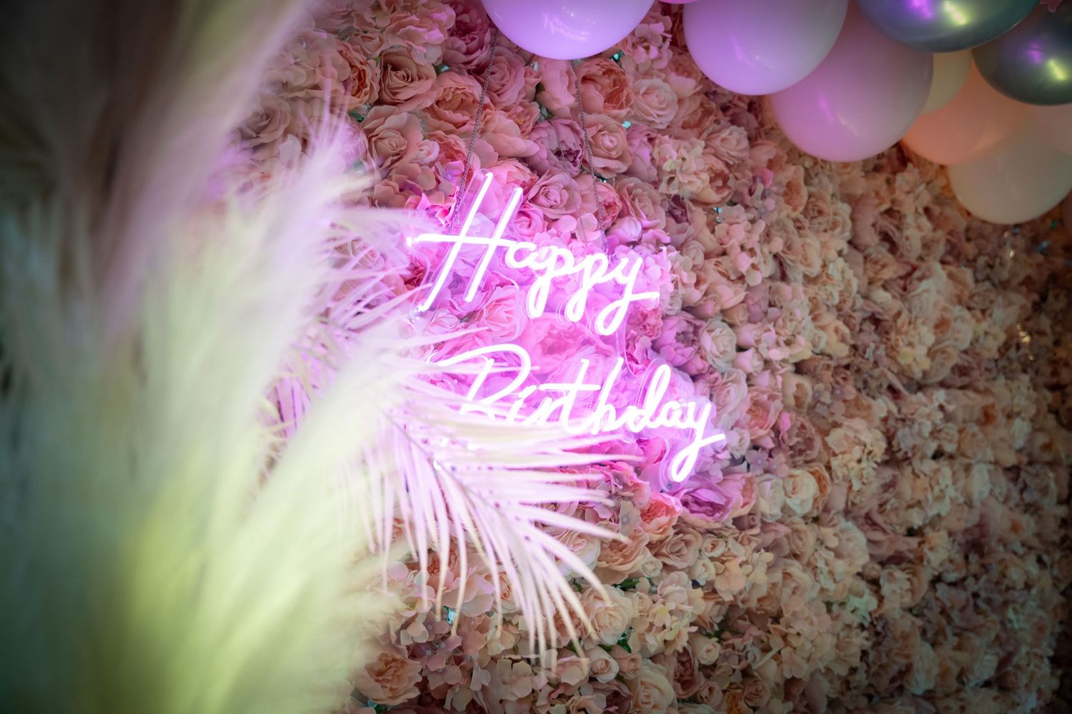 Pink neon 'Happy Birthday' sign glowing against floral wall with palm leaf shadow.