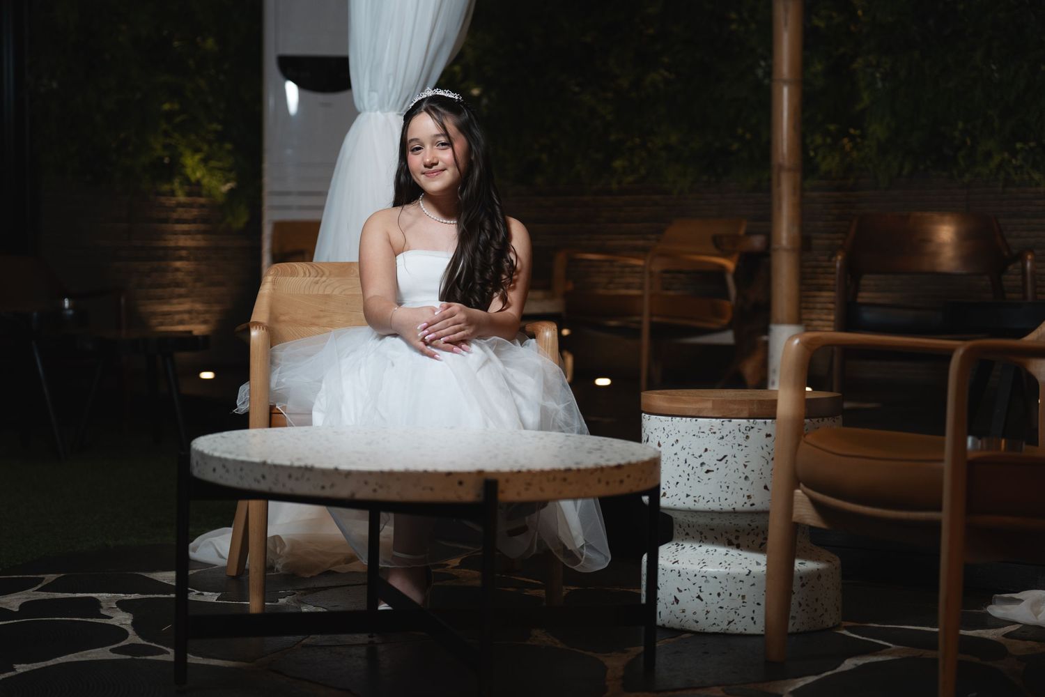 A bride in a white wedding dress sits on a rustic wooden bench in a dimly lit venue setting.