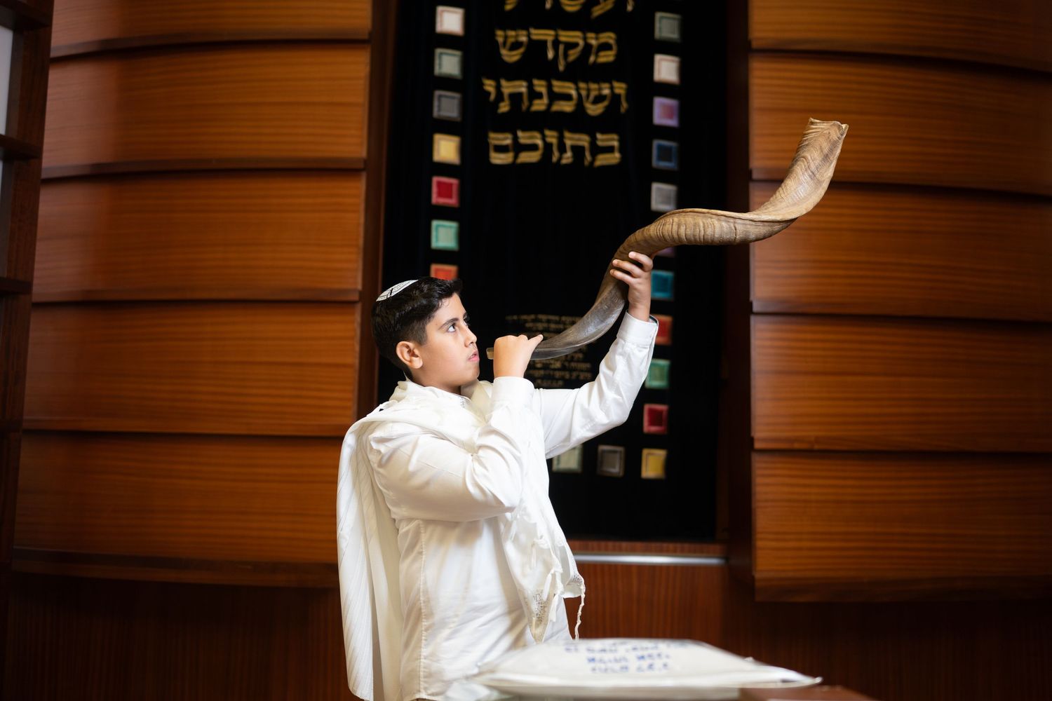 Person in white garment blowing shofar in front of wooden synagogue wall with Hebrew text.